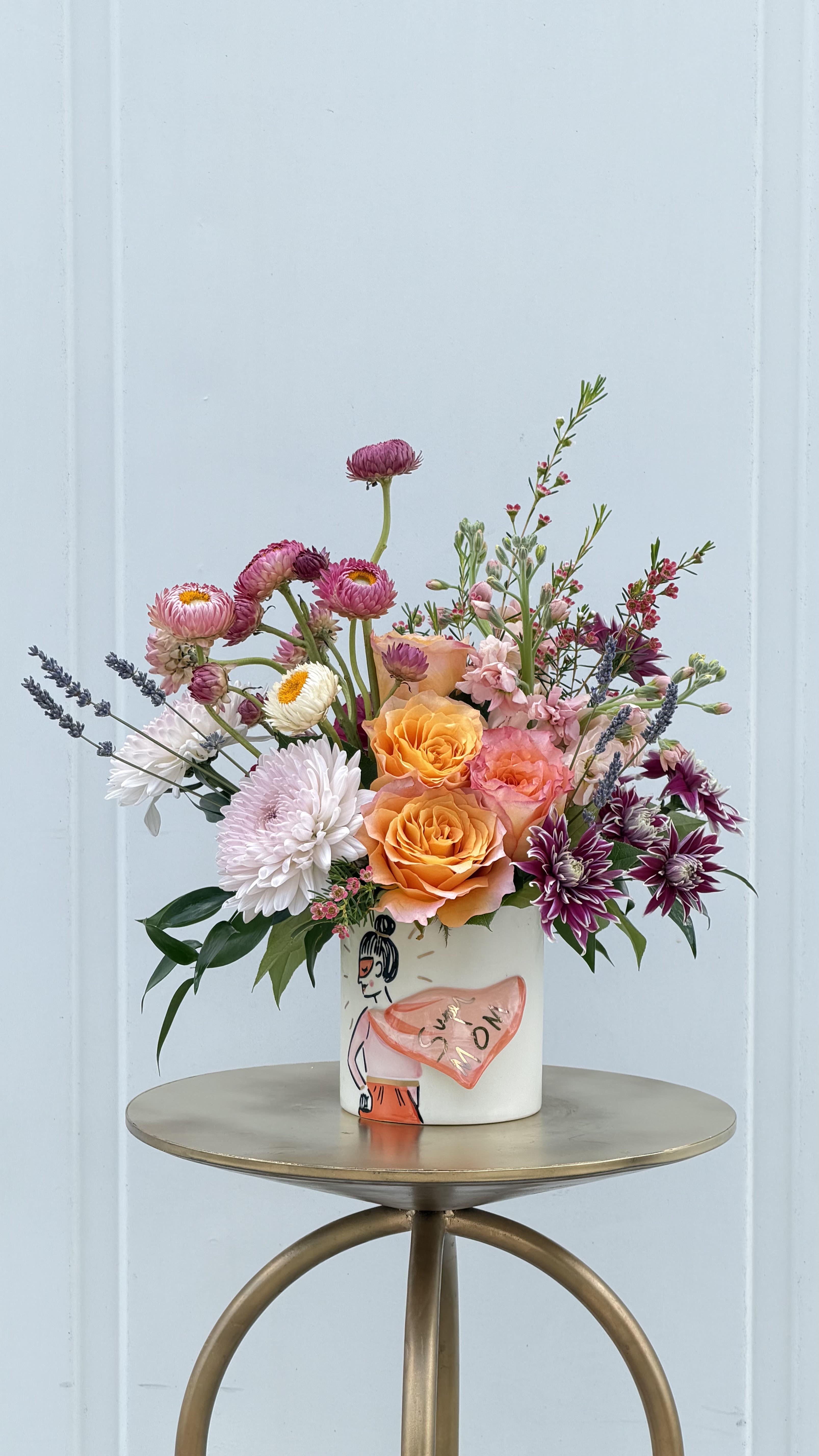 Super Mom - Celebrate her with a bouquet as vibrant and beautiful as she is. This Mother’s Day arrangement features glowing peach and coral roses surrounded by soft blush blooms, delicate pink accents, and airy seasonal florals for a fresh, garden-inspired look.  Designed in a charming ceramic keepsake vase with a sweet “Super Mom” message, it’s more than just flowers—it’s a heartfelt reminder of how much she’s loved and appreciated.  Perfect for brightening her home and her day, this arrangement is a thoughtful way to say thank you for everything she does.  ***Limited quantity available***