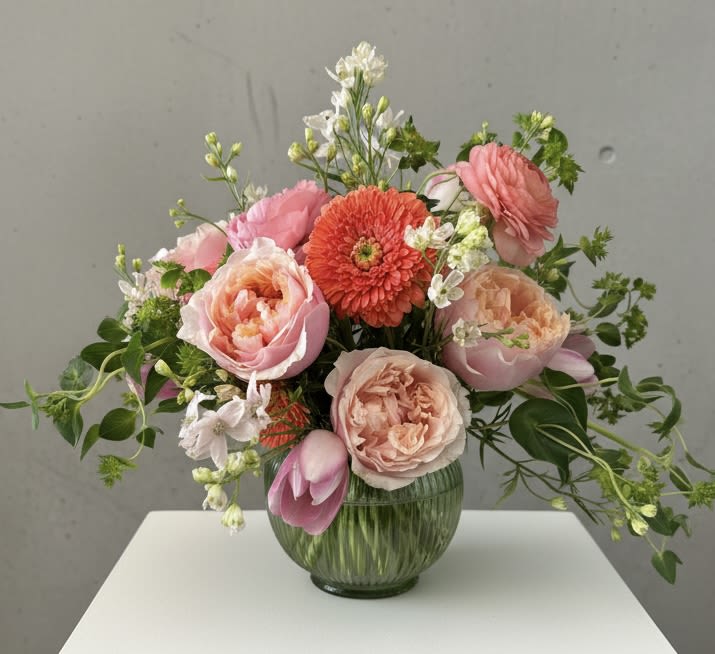 Momosa - Soft, garden-fresh pinks and peach tones mingle in this Momosa flower arrangement-perfect as a birthday bouquet, Mother's Day gift, or just-because surprise for someone who loves a feminine, lightly celebratory touch. The mix of garden/standard roses, ranunculus ,coral gerbera daisies, and pink tulips reads warm and affectionate; a few white accent blooms and green foliage lift the palette so the recipient feels seen and joyful.  Arranged loosely in a ribbed glass vase, the design is airy and hand-gathered, with stems and small filler blooms extending outward for an organic, garden-picked look. Expect a rounded composition roughly suited for tabletops or a kitchen counter; the textured vase adds subtle vintage charm while keeping the focus on the soft florals.  This is the kind of bouquet that makes a mom, friend, or partner feel genuinely celebrated-cheerful without being over-the-top. Vase-inclusive and ready to display on arrival; refresh water every 2-3 days and trim stems slightly to extend bloom life.