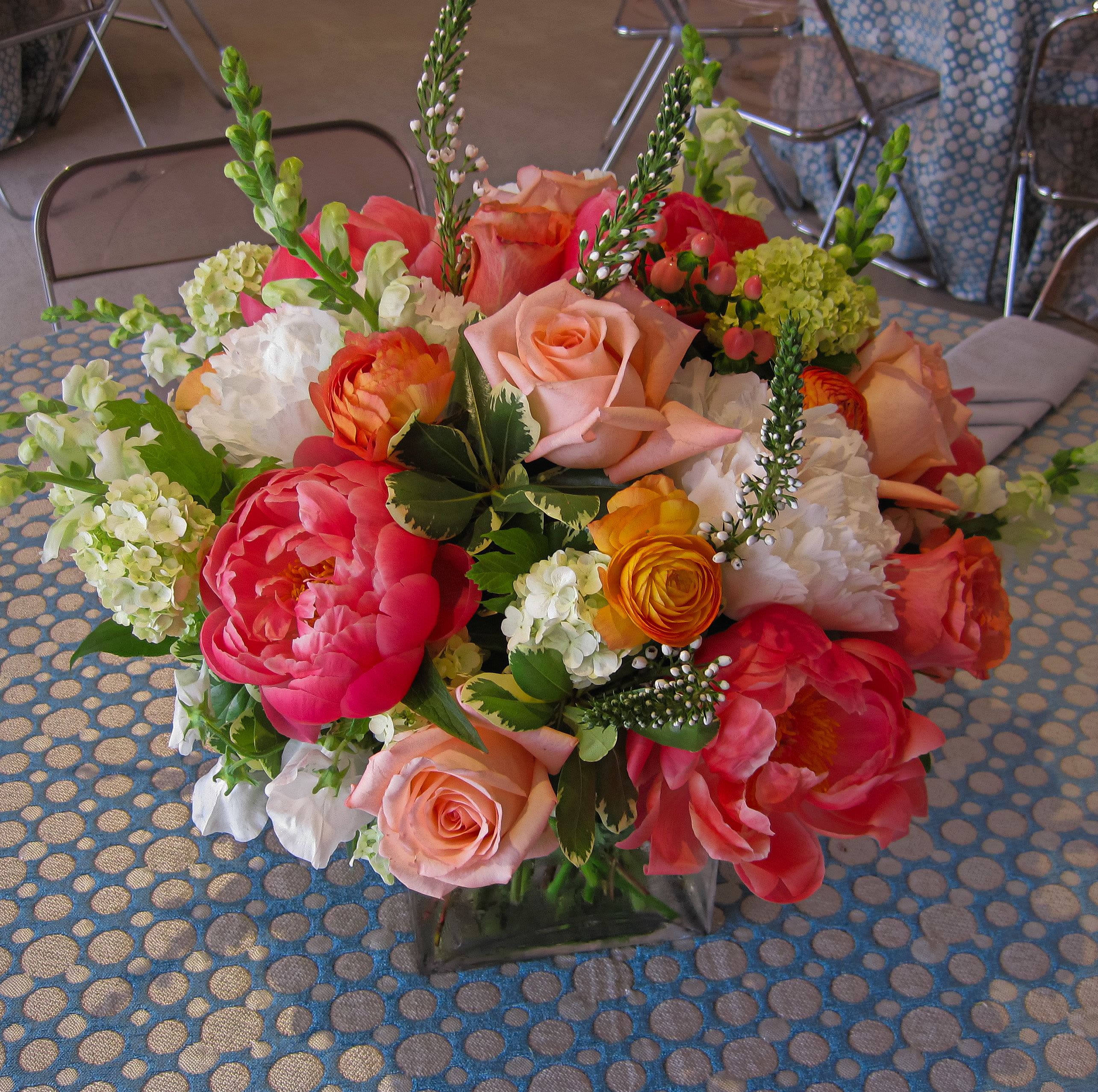 Low arrangement of pink peonies, peach roses, and white blooms in a clear glass cube vase