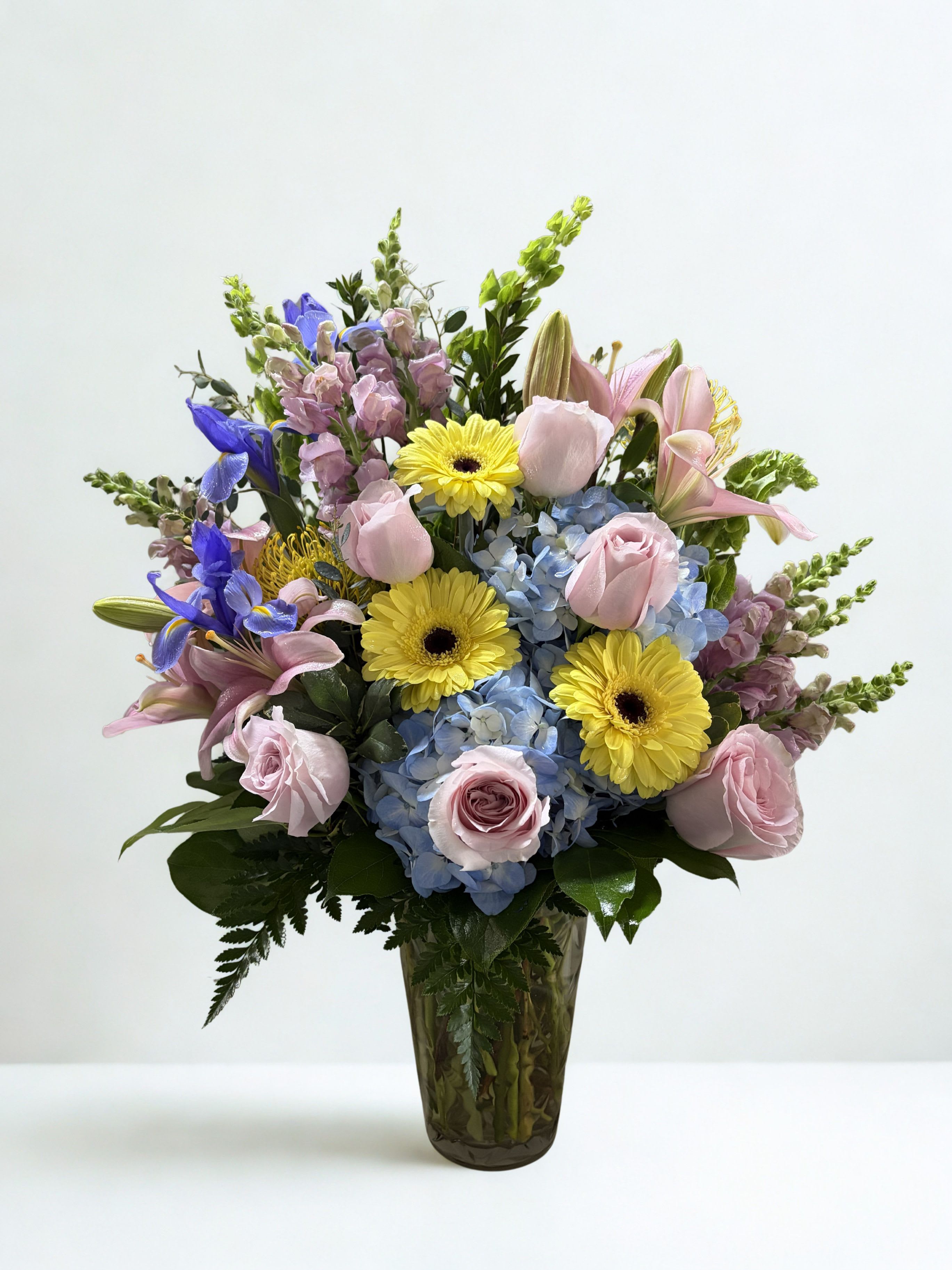 Soft Skies - Soft pink roses and cloud-like blue hydrangeas mingle with bright yellow gerbera daisies and lavender-purple accents in a garden-style bouquet that feels like a breath of fresh spring air. This flower arrangement is ideal as a birthday bouquet, a thank-you gift, or a thinking-of-you surprise-they'll feel genuinely lifted by the soft, cheerful color story and the graceful lilies and snapdragons that add gentle height and movement.  Arranged in a clear glass vase, the design is full and rounded at the base with taller lily blooms and snapdragon spikes rising above for a layered, natural look. Textures range from the velvety rose petals and ruffly hydrangea clusters to the crisp faces of the gerbera daisies and the delicate snapdragon stems, creating visual depth and a joyful pastel palette of pink, blue, yellow, and purple.  Perfect for spring celebrations or everyday brightening, this bouquet arrives ready to display and makes a thoughtful, uplifting gift for the person you're thinking of. For best enjoyment, place in a cool spot out of direct sun and replenish vase water every two days to keep blooms looking fresh.