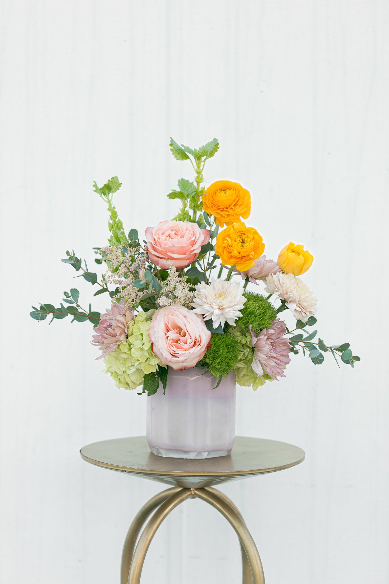 Sweet Treat - Sweet Treat is a cheerful, garden-inspired arrangement bursting with soft pastels and playful pops of color. Blush roses and delicate chrysanthemums create a romantic foundation, while vibrant golden ranunculus add a sunny, uplifting touch. Accents of fresh greenery and textured blooms bring depth and movement, giving the design a naturally gathered, just-picked feel.  Arranged in a soft blush glass vessel, Sweet Treat is perfectly sized for a desk, bedside table, or small gathering space—anywhere that could use a little extra joy. Its light, airy palette and whimsical charm make it an ideal gift for celebrations, thank-yous, or simply brightening someone’s day.  Fresh, sweet, and full of personality—Sweet Treat is happiness in bloom.