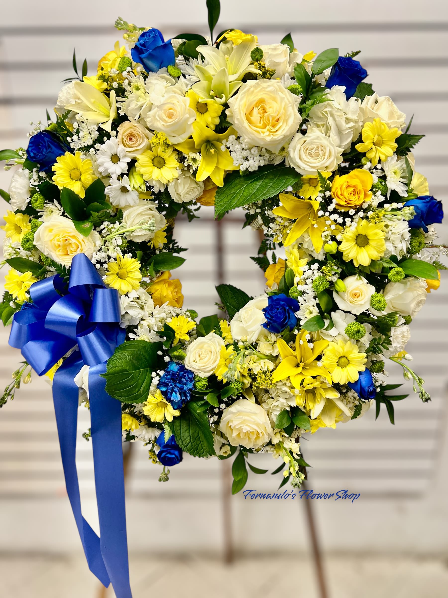 Standing wreath of white, yellow, and blue flowers with a large blue bow on an easel.