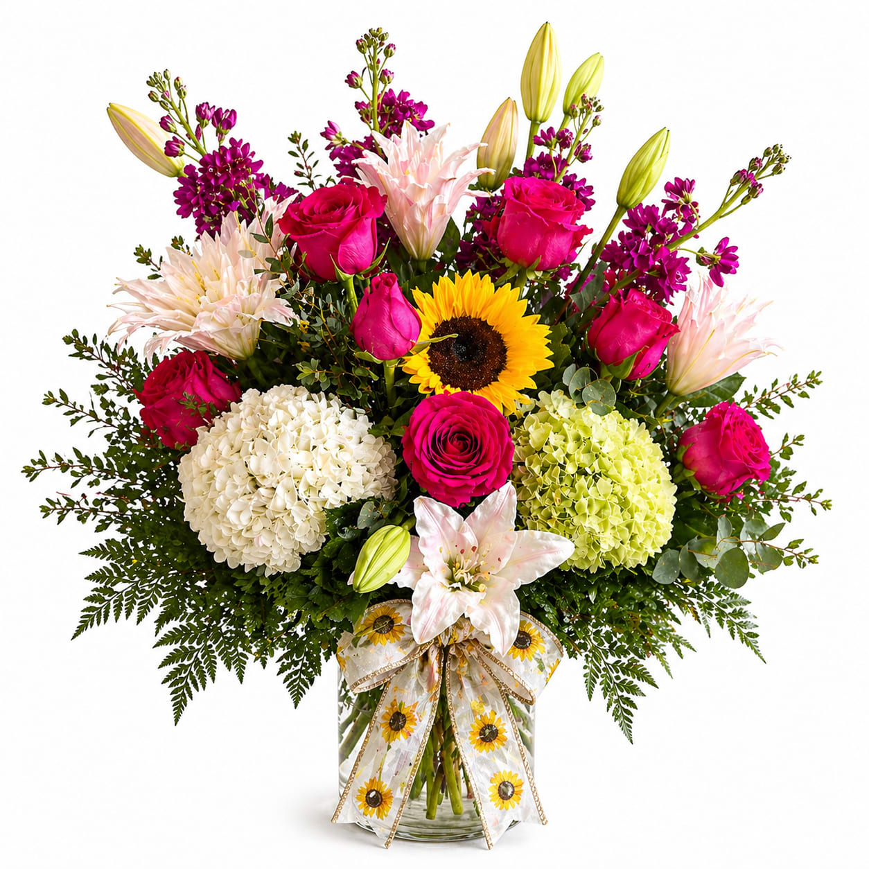 A Mother’s Love - Bright, layered and joyfully colorful, this large mixed flower arrangement-perfect as a birthday bouquet, Mother's Day gift, or cheerful thank-you-is built to make her feel seen and celebrated. Hot-pink roses and pale pink lilies form the vibrant heart, while a single bright sunflower at the center and purple filler blooms add surprise and sunny optimism. The overall effect reads lively and generous; it announces the occasion with confident color and a warm, personal touch.  Arranged in a clear glass vase and tied with a sunflower-patterned decorative ribbon, the design pairs white and lime-green hydrangea with creamier tones for a full, rounded silhouette accented by taller buds and branching stems for height. Fern-like greenery frames the base, giving the bouquet a textured, garden-fresh look. This presentation is offered as a gift-ready set with a box of Russell Stover pecan delights included, making it an easy, celebratory choice for moms, friends, or anyone who deserves a bright, uplifting surprise.