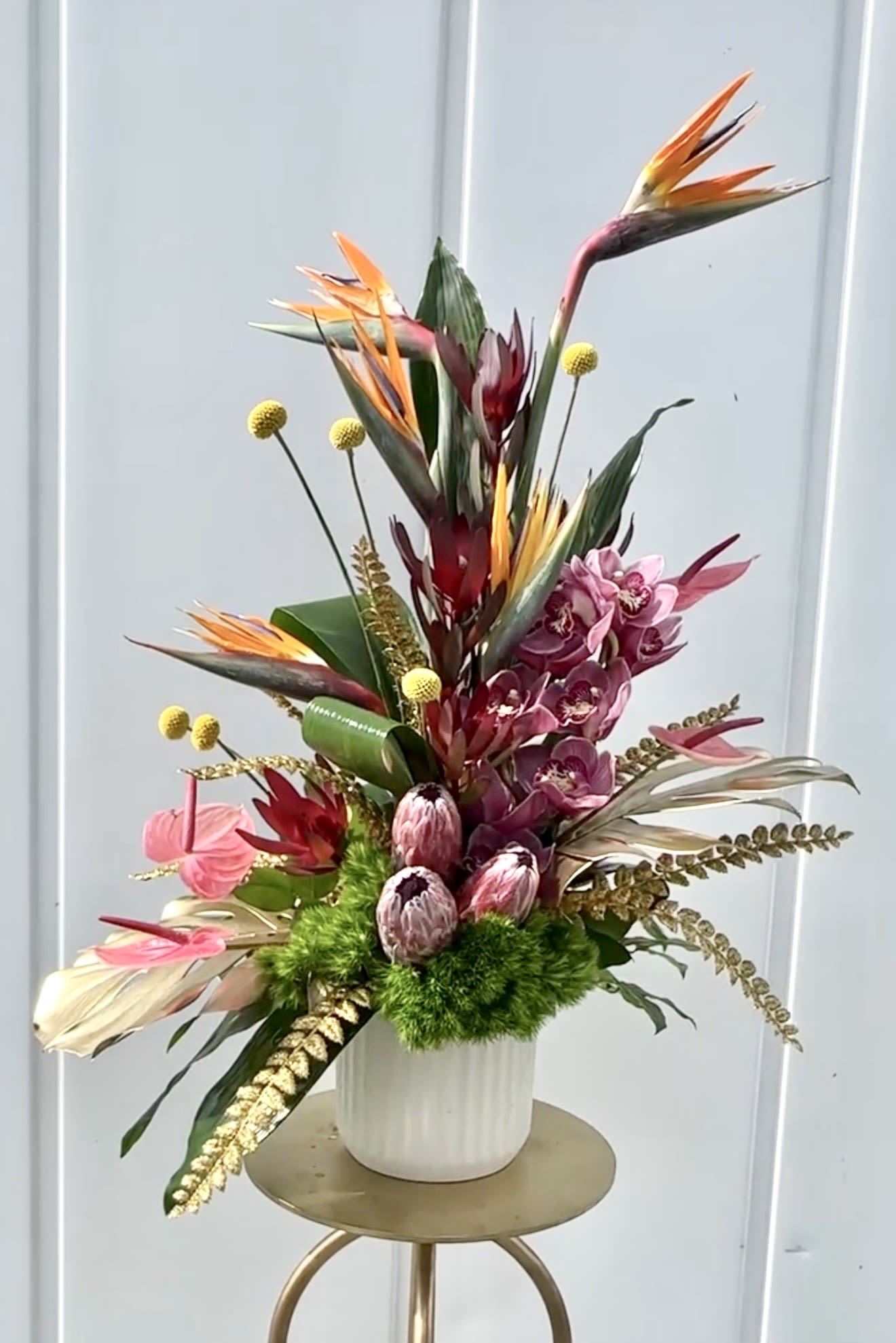 Madre Mia - Madre Mia is a bold, expressive arrangement that radiates passion and vibrant energy. Striking birds of paradise reach upward with dramatic flair, complemented by rich pink orchids and sculptural tropical blooms that create depth and movement. Accents of golden textures, playful billy balls, and lush greenery add a dynamic, layered feel that captures attention from every angle.  Designed in a modern ceramic vessel, Madre Mia blends exotic beauty with artistic composition, making it a true statement piece. Perfect for celebrations, grand gestures, or elevating any space with a touch of the unexpected, it brings warmth, color, and personality wherever it’s placed.  Daring, lively, and unforgettable—Madre Mia is a celebration of bold beauty and joyful expression.