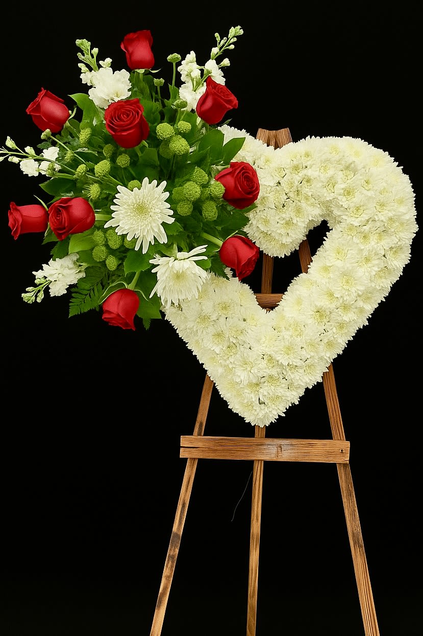 Heart-shaped white floral wreath on an easel beside a red rose bouquet