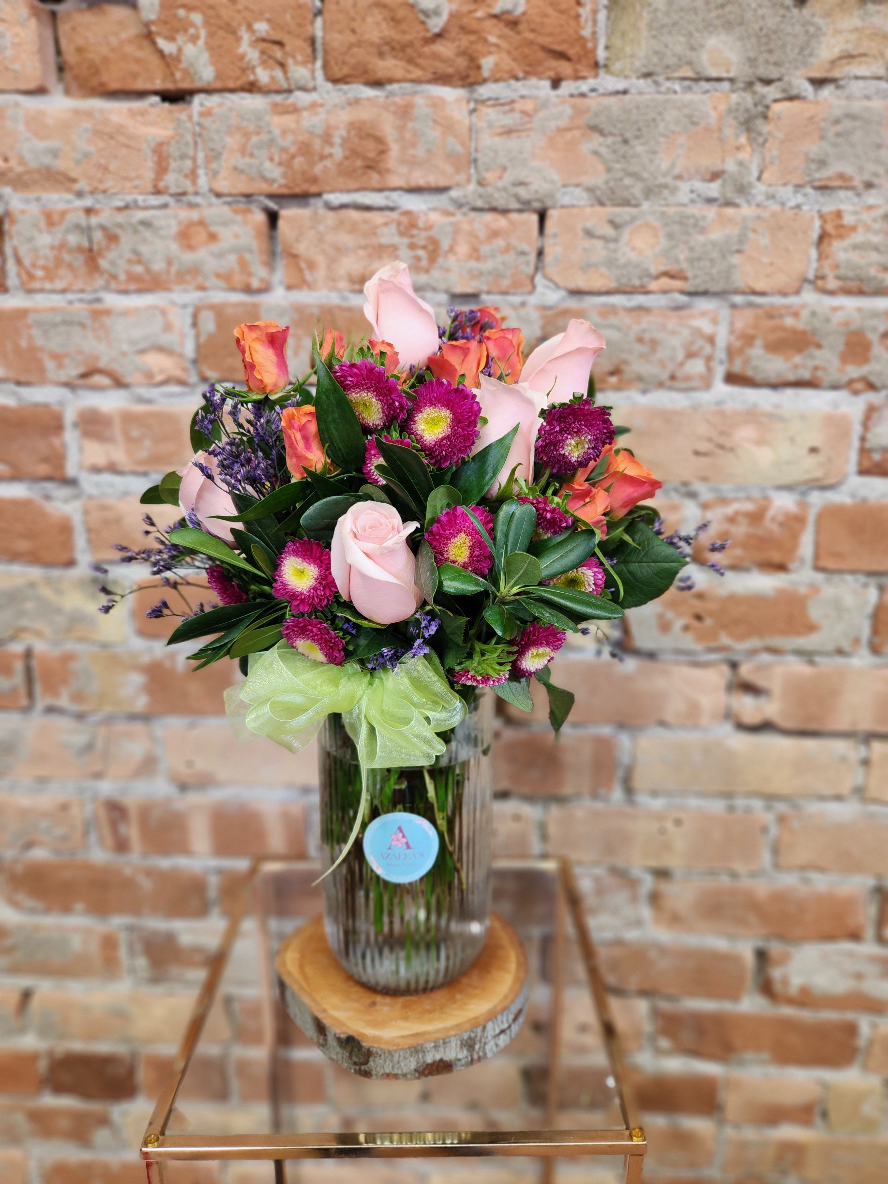 Pink roses and purple flowers arranged in a glass vase with a green ribbon.