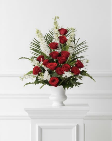 Red and White Urn - Multiple red roses with white filler and palm-like greenery, arranged upright in a white ceramic vase.
