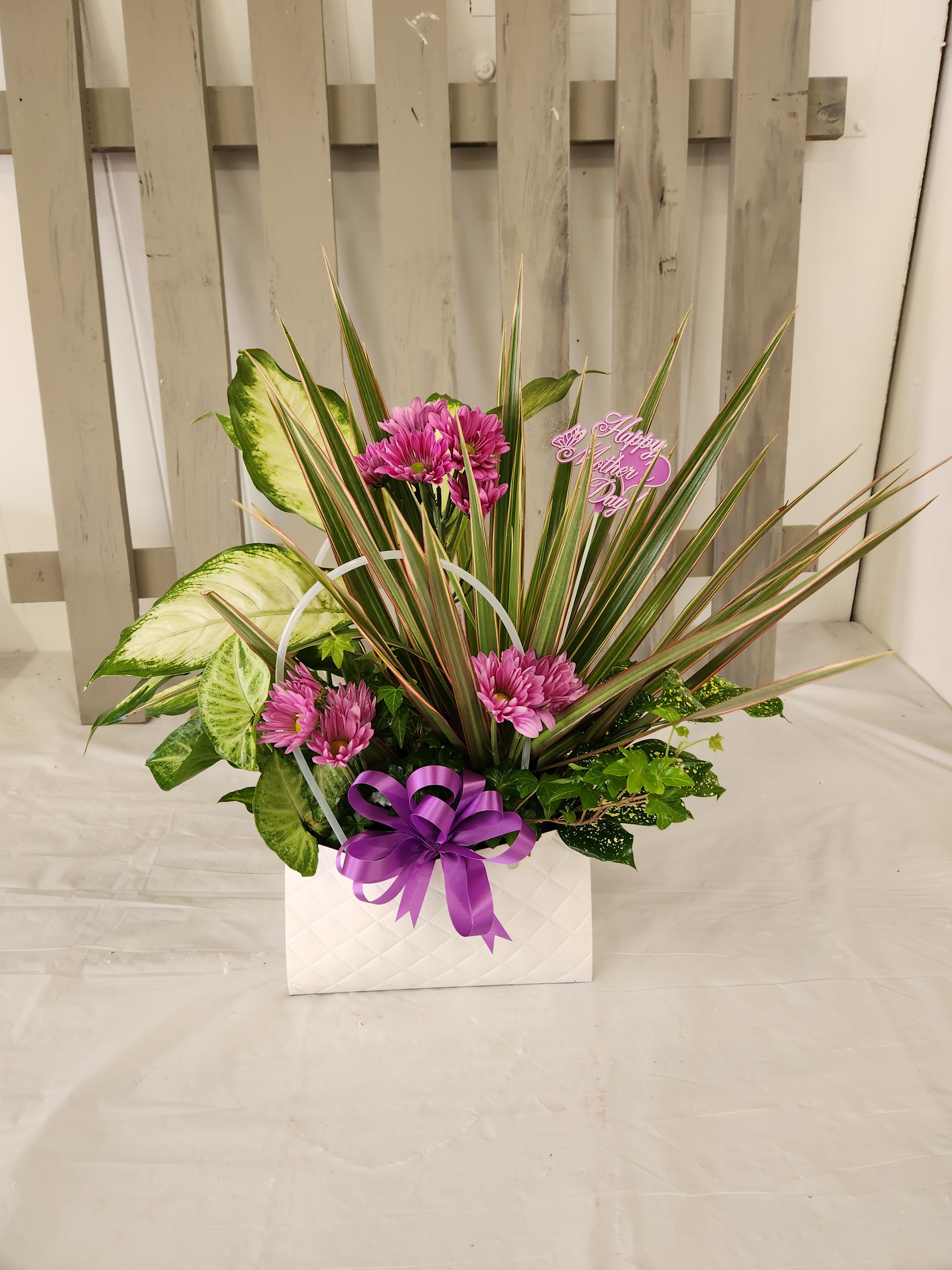 White Purse - Bright pink daisy-like blooms and dramatic variegated green foliage create a cheerful, textured centerpiece that makes someone feel genuinely seen. This flower arrangement for Just Because or Thank You moments is presented in a white quilted gift box and finished with a large purple ribbon and a small "Happy Mother Day" pick, giving it a celebratory, slightly tropical feel perfect for Mother's Day or everyday appreciation.  The mix pairs soft round blossoms with long pointed leaves for bold contrast-pink, purple, white and green tones play together for a lively, feminine palette. Arranged compactly in the white gift box, the display is finished with a discreet plastic floral pick and sits at an easy-to-place size ideal for tabletops, desks, or kitchen counters. It's the kind of thoughtful surprise that brightens a room and lets the recipient know you're thinking of them.