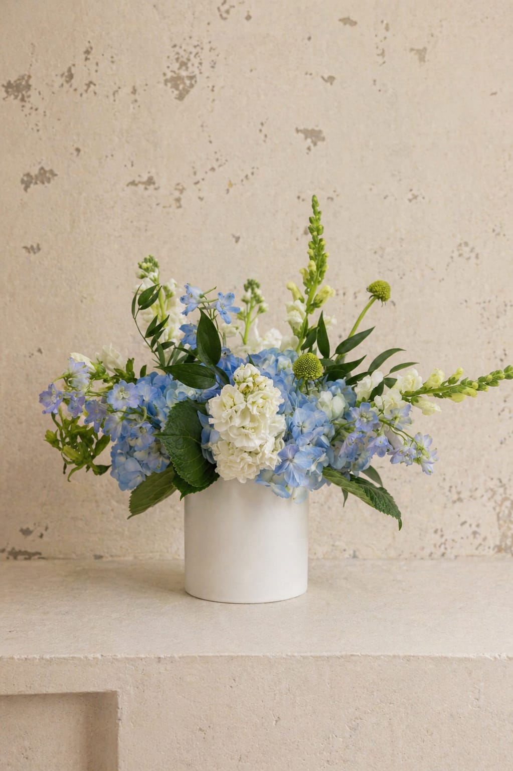 The Keepsake - A soft, garden-inspired arrangement designed to feel as special as she is. This piece features lush blue hydrangea, delicate delphinium, creamy white blooms, and fresh seasonal greens, thoughtfully arranged in a modern white ceramic vessel.  Airy, elegant, and naturally beautiful, it’s the perfect way to celebrate Mother’s Day, whether for a quiet moment at home or a thoughtful gesture she won’t forget.  Each arrangement is handcrafted with the freshest blooms available, so slight variations may occur while maintaining the overall look and feel.
