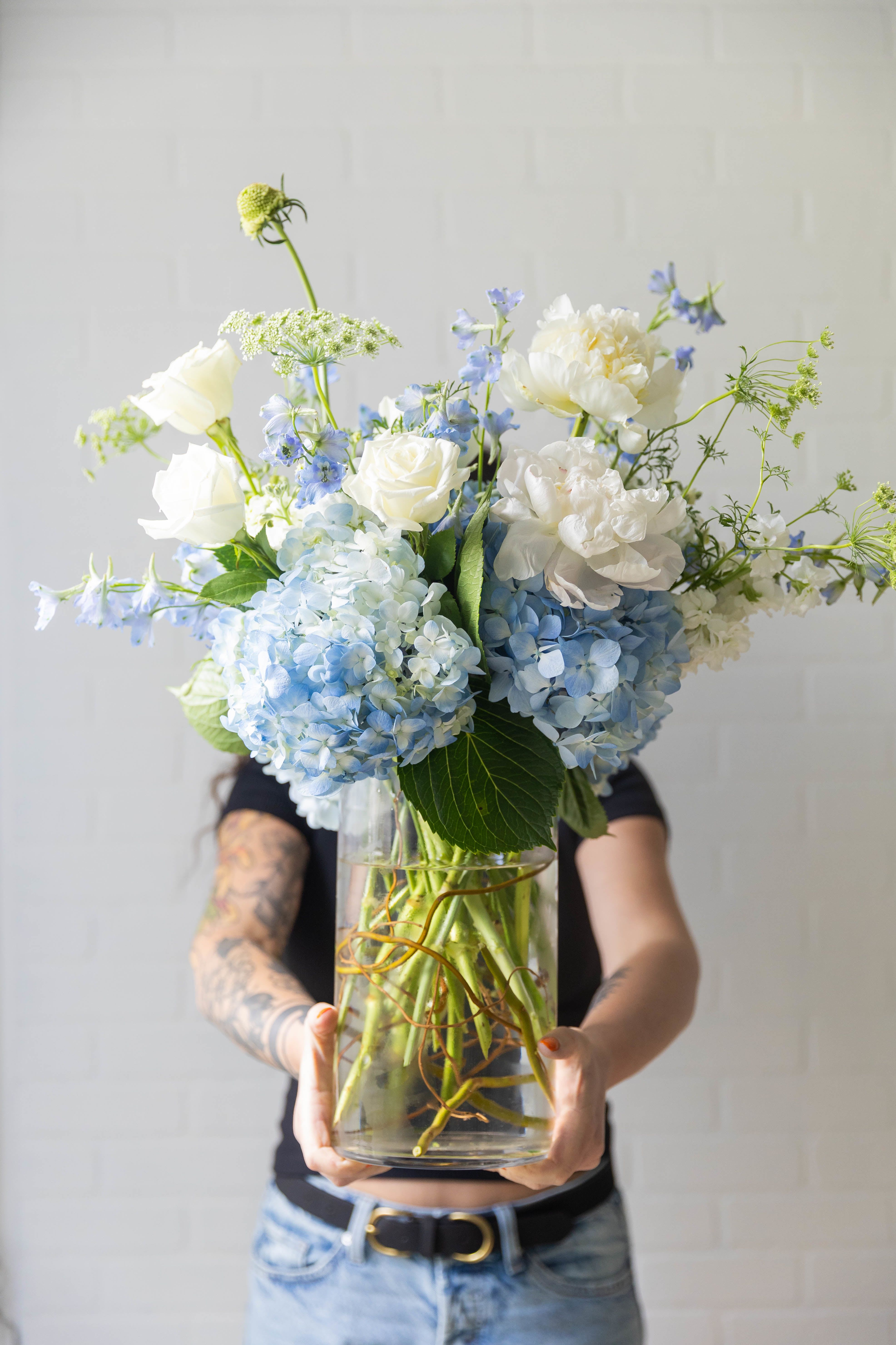 Fleur de Mere - A soft, garden-inspired arrangement in calming blue and ivory tones, featuring lush hydrangea, delicate delphinium, and romantic seasonal blooms. Designed in a large clear glass vase for an effortless, natural look, perfect for Mother’s Day. 
