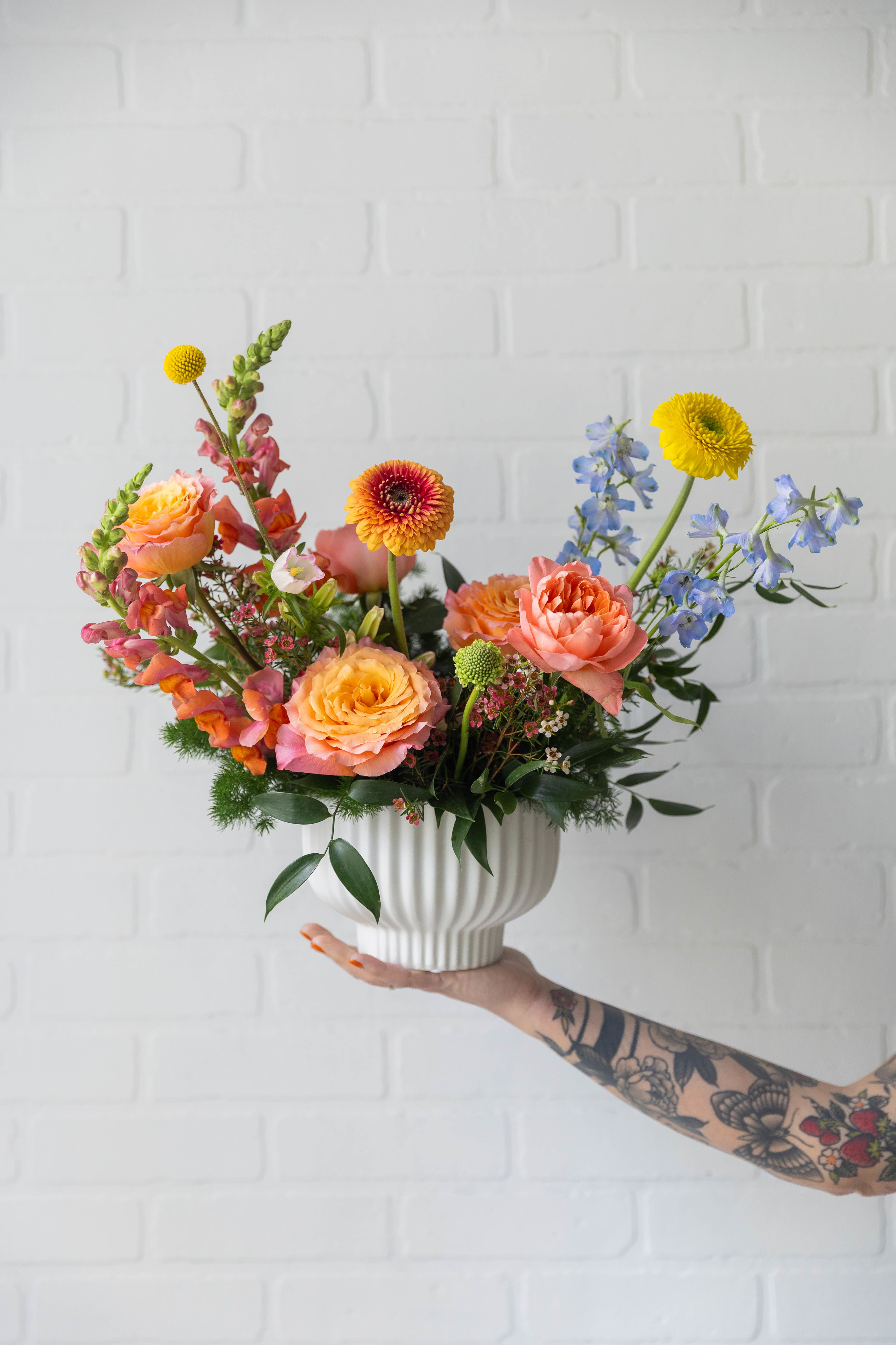 Where Love Grows - A joyful, garden-inspired arrangement in warm peach, coral, and golden tones, accented with soft blues and fresh greenery. Designed in a textured ceramic vessel, it’s a vibrant and heartfelt way to celebrate Mom.