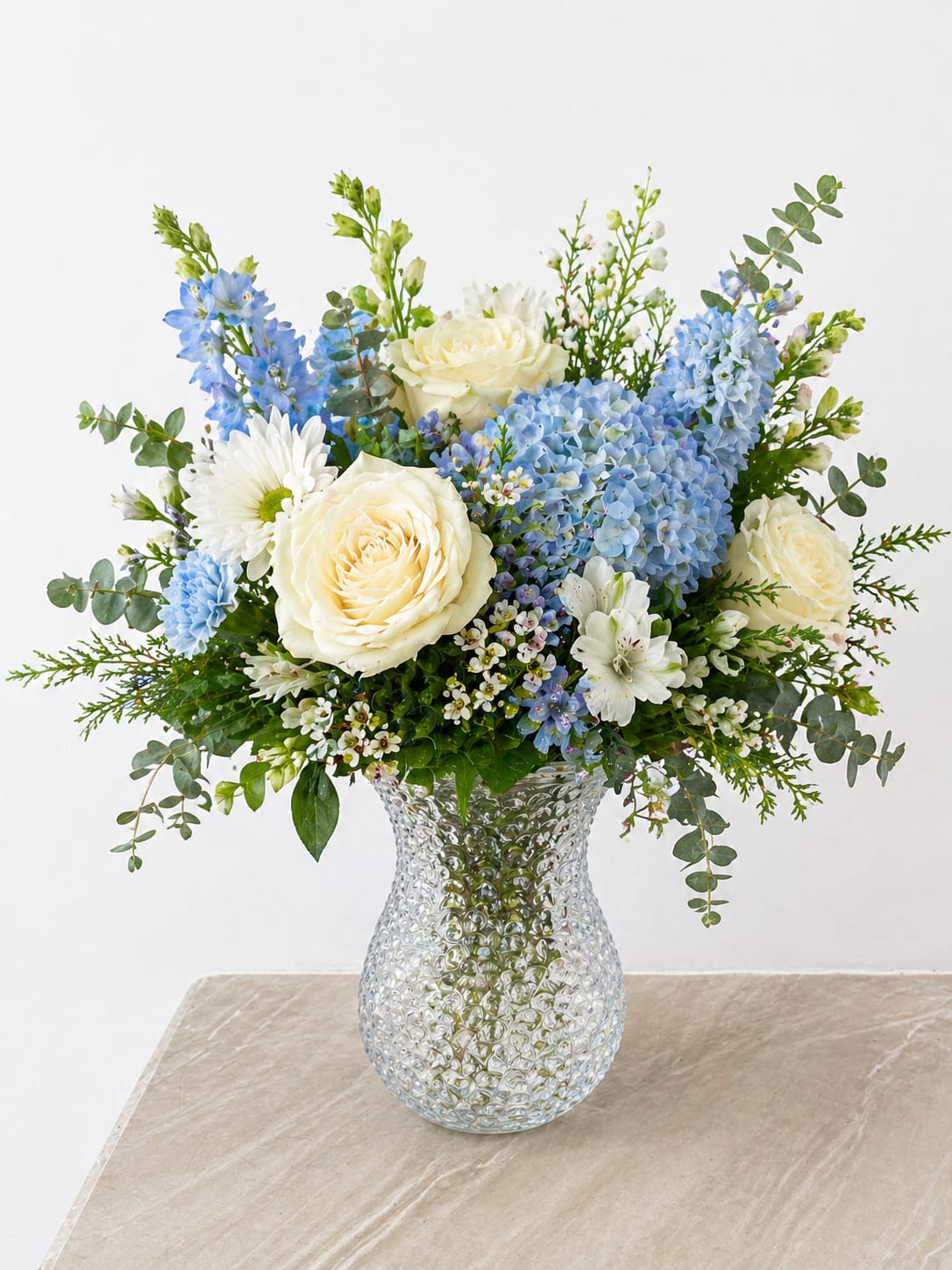 Blue &amp; White Garden Vase Arrangement - Soft, fresh, and effortlessly elegant, this arrangement features a beautiful blend of cool blue and crisp white blooms arranged in a textured glass vase. Lush hydrangea, creamy white roses, and delicate seasonal flowers are thoughtfully paired with airy greenery to create a full, garden-inspired design.  The mix of textures and soft tones brings a calming, sophisticated feel—perfect for sympathy, celebrations, or simply brightening a space. The clear patterned vase adds a subtle touch of charm while allowing the natural beauty of each stem to shine.  Handcrafted with care, every arrangement is uniquely designed to feel balanced, fresh, and naturally beautiful.