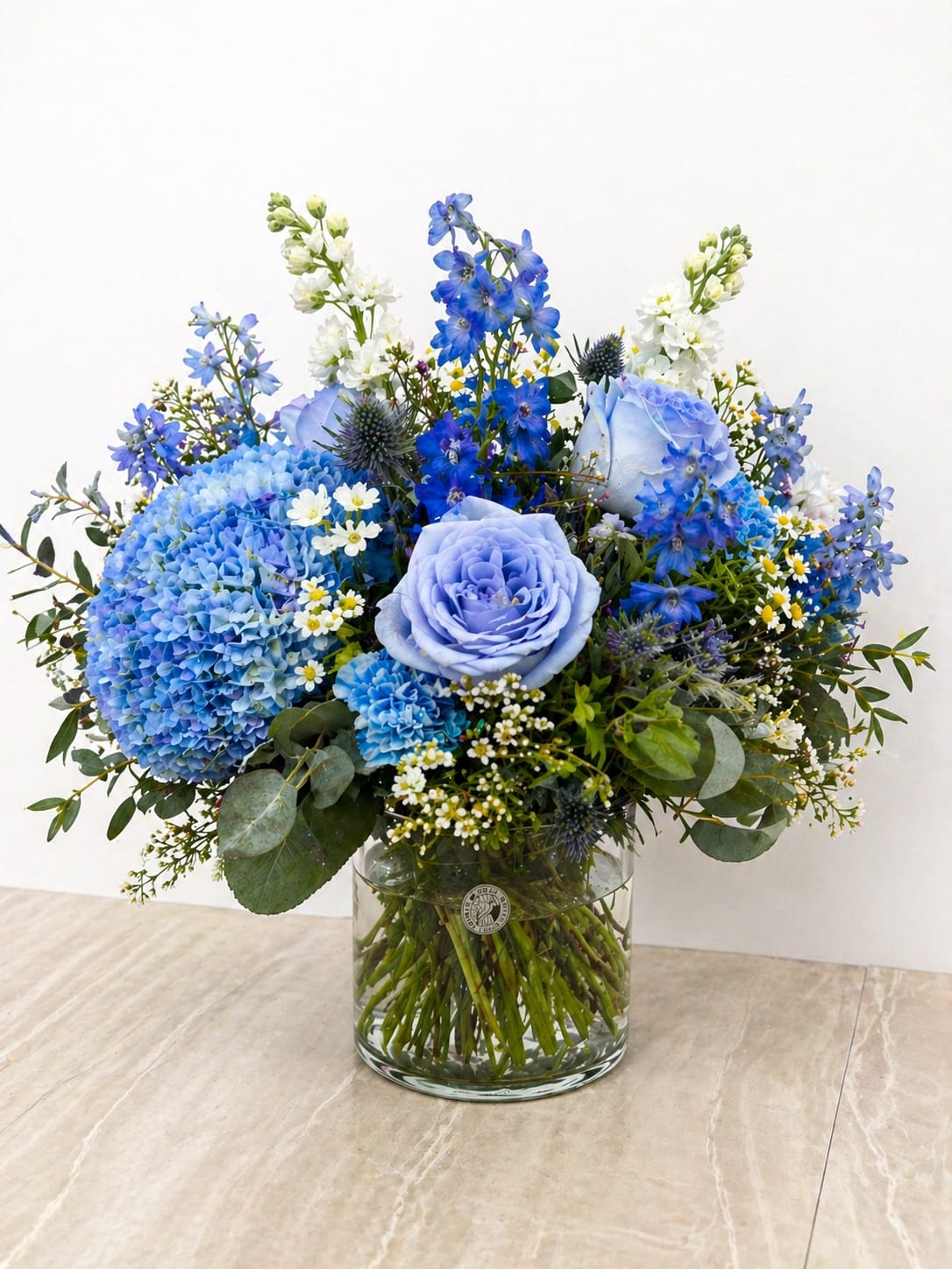 Full of Radiance  - TThis elegant floral arrangement features a refreshing blend of soft blue and crisp white blooms, accented with delicate touches of yellow and lush greenery. Designed in a clear glass vase, the composition showcases premium flowers such as hydrangea, seasonal fillers, and roses that are artfully enhanced with a soft blue floral tint for a beautifully cohesive look.  The gently tinted roses add a unique, modern touch while maintaining a natural, garden-inspired feel. Full of texture and airy movement, this arrangement brings a sense of calm and sophistication to any space.  Perfect for celebrations, its cool color palette makes it both timeless and versatile for any occasion or décor style.  Handcrafted with care, each design is one-of-a-kind and thoughtfully arranged to highlight the beauty of every stem.   