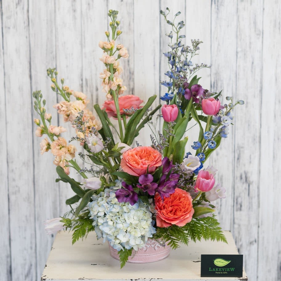 Skylark - A whimsical, garden-style flower arrangement that feels like a breath of spring - Skylark is a mixed-color Mother's Day arrangement built to make her smile. Tall, airy stems rise above a fuller cluster of blooms in a soft pink container, combining pink tulips, coral garden roses, pale blue hydrangea, delphinium and stock with purple accents for a fresh, romantic look that reads as both celebratory and thoughtful.  Hand-arranged in a low pink container, Skylark blends texture and movement: the hydrangea provides a lush base, tulips and roses bring graceful curves, and the delphinium and stock add height and vertical interest. Colors include pink, blue, purple, peach and white for a palette that is perfect for telling Mom she's cherished.  Approx. 16"H x 14"W *This item is a Mother's Day Special and is available May 7-9th*