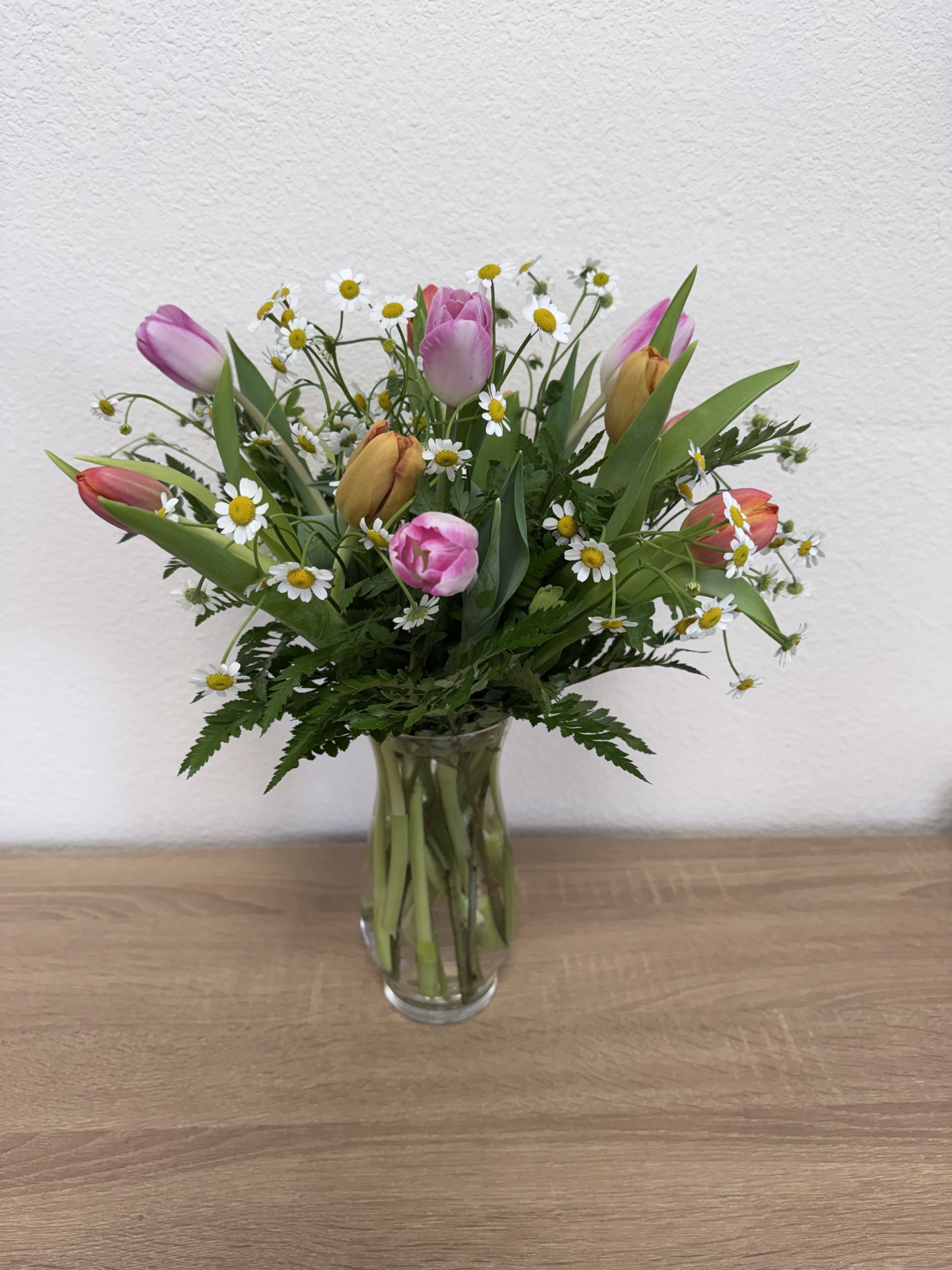 Tulips for Mom - Bright tulips mingle with fever- few fillers in a clear glass vase for a fresh, garden-style bouquet perfect for Mother's Day. This flower arrangement - perfect as a thank-you or just-because surprise for Mom - reads as cheerful and springlike, the airy spacing giving it a light, casual feel that will make the person you're thinking of feel genuinely seen.  One dozen tulips are loosely gathered, letting stems and greenery breathe for that wildflower look. Presented in a simple clear glass vase, the arrangement stands at a natural, relaxed height and showcases pink, white, yellow, and green tones that brighten a kitchen table or bedside. change water every two days and trim stems slightly to keep blooms fresh.