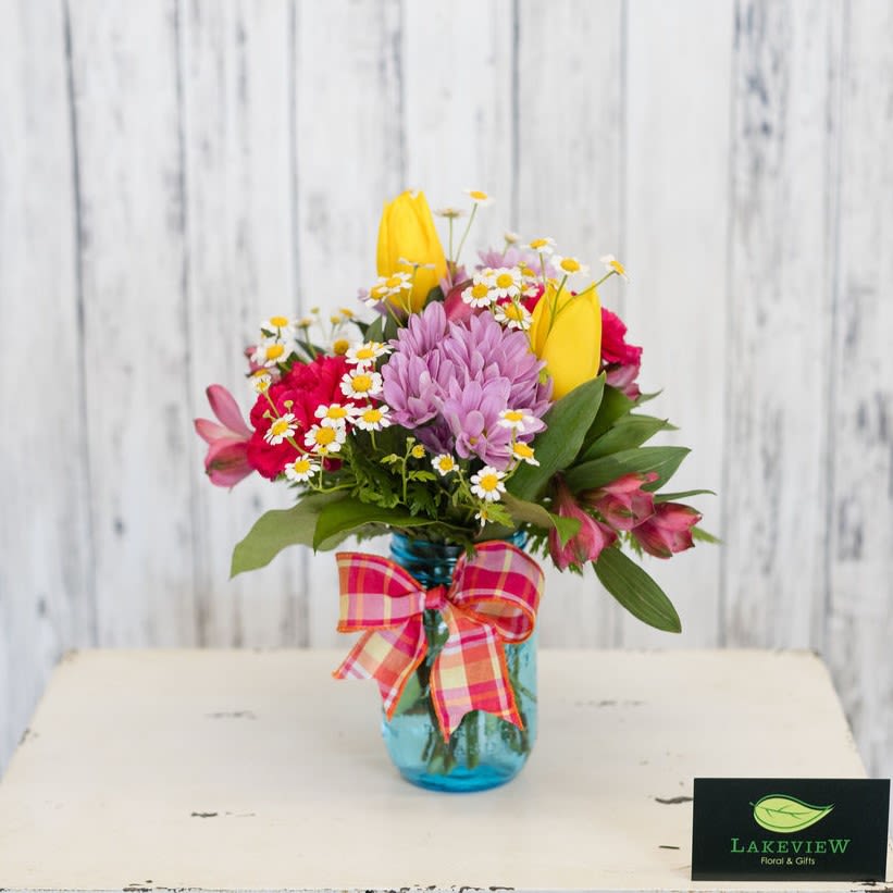Bespoke - A bright, garden-style flower arrangement designed to make her feel genuinely celebrated-perfect as a Mother's Day gift. This mixed bouquet pairs playful-pink, peach, yellow, white and soft purple blooms-with a casual, handcrafted look that reads fresh and joyful the moment it arrives.  Presented in a petite blue mason jar and finished with a pink-and-orange plaid ribbon, blooms include carnations, tulips, alstroemeria, daisies and chamomile arranged in a loose, slightly rustic style. The composition mixes saturated yellow tulips and magenta carnations with lavender chrysanthemums and white daisy sprays for lively contrast; overall size is about 8"H x 5"W.  Thoughtfully sized for a desk, bedside table, or kitchen counter, this arrangement is the kind of gift that brightens someone's day and makes them feel celebrated the moment they receive it!  Approx. 8"H x 5"W *This item is a Mother's Day Special and is available May 7-9th*
