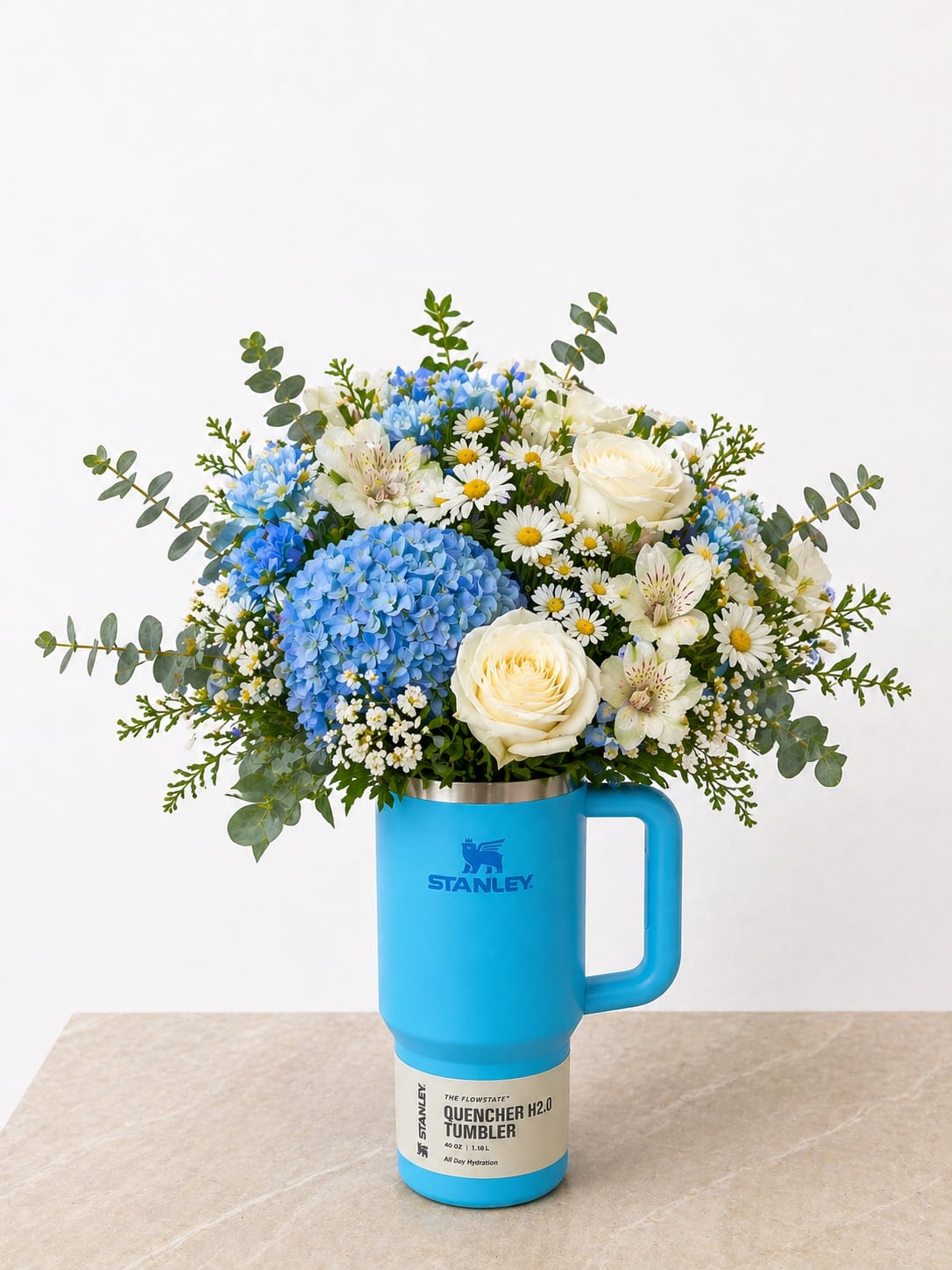 Blue Stanley Floral Arrangement - A fresh and modern twist on a classic gift, this design features a vibrant blue Stanley tumbler paired with a lush, hand-crafted floral arrangement. Filled with soft blue hydrangea, creamy white roses, delicate daisies, and touches of fresh greenery, this piece brings together beauty and everyday functionality in one thoughtful design.  The cheerful mix of textures and colors creates a bright, airy feel, while the reusable Stanley tumbler adds lasting value long after the flowers are enjoyed. Perfect for birthdays, celebrations, or just because, this arrangement is both stylish and practical—making it a gift they’ll truly love and use.  Each arrangement is uniquely made with care, ensuring a full, balanced look that highlights every bloom.