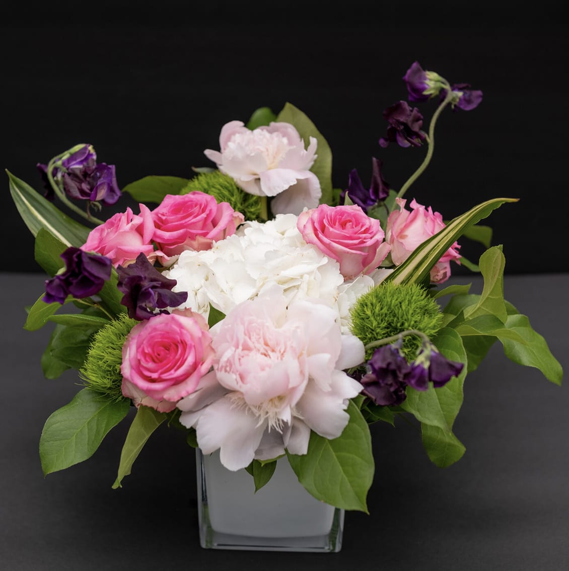 Delight Lovely Spring - Mother's Day - Bright pink roses and soft white blooms create a joyful, romantic statement in this springtime flower arrangement-perfect as a Mother's Day bouquet, birthday surprise, or thank-you gift for someone who loves classic color harmony. The compact, layered shape reads fresh and modern, so the person you're celebrating will feel seen and appreciated the moment the vase arrives.  This arrangement pairs vivid pink roses with delicate white flowers, purple accent blooms, and green foliage for contrast. Set in a clean square glass vase, the design mixes full-petal roses and carnation-like textures with airy trailing stems to give depth without appearing fussy. The balanced, rounded silhouette fits well on a mantel, bedside table, or office desk.  Choose this for a mom, friend, or colleague who enjoys feminine, cheerful florals-it's the kind of bouquet that brightens a room and lifts spirits. Arrives ready to display in the included square glass vase; simple water care and fresh-cut stems will keep these blooms looking their best.