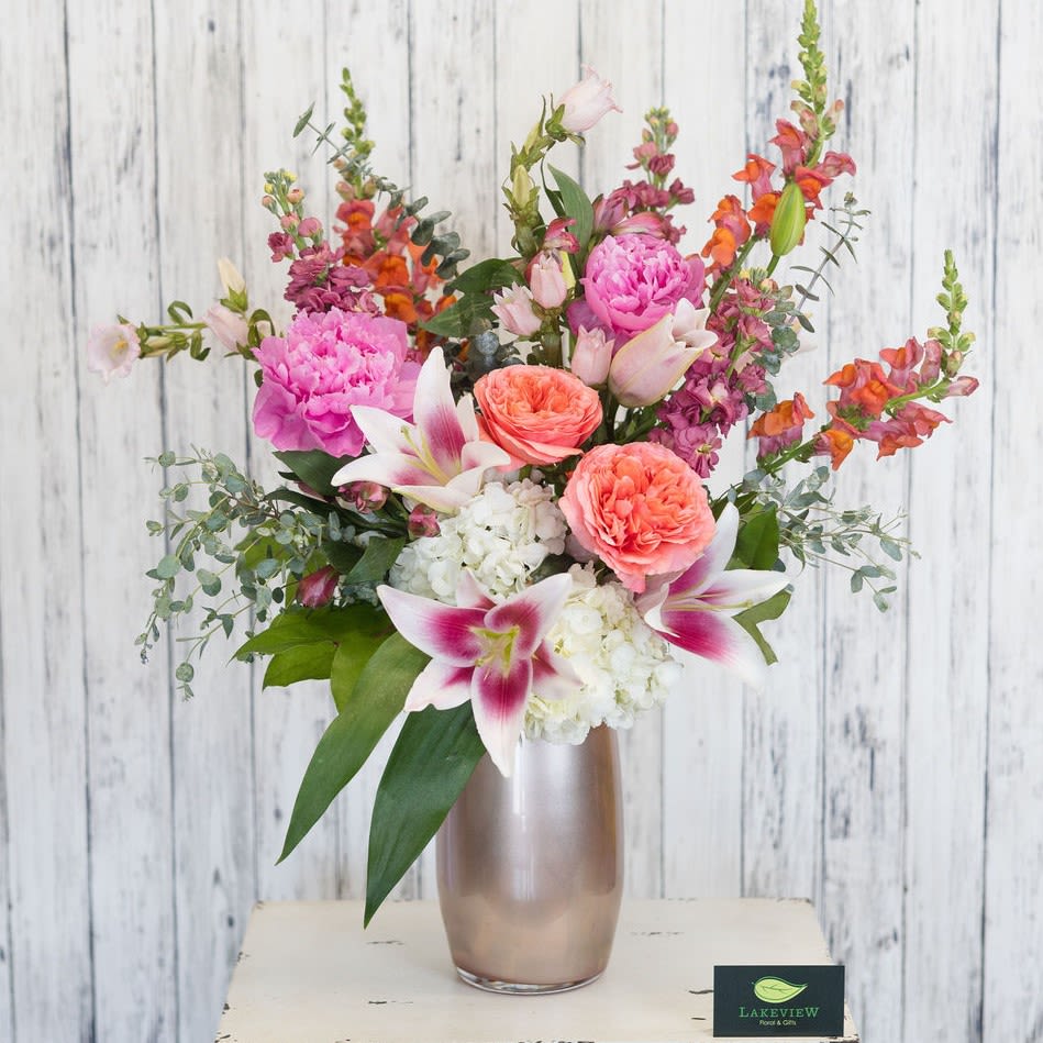 Nightingale - A striking, garden-style arrangement bursting with peach, orange, pink, white and touches of magenta is arranged upright in a tall metallic vase-designed to make the Mom you're thinking of feel celebrated and seen. This flower arrangement for Mother's Day  greets the room with a lively, romantic presence: tall snapdragon spikes and campanula stems give movement while rounded peonies and hydrangea soften the silhouette.  Filled with garden roses, peonies, stock, snapdragons, lilies, hydrangea, alstroemeria and campanula, the mix balances ruffled textures and sculpted blooms in coral, blush, bright pink, white and green foliage. The smooth metallic vase adds a polished, contemporary finish that complements the airy, layered composition; the overall design reads as both lush and effortlessly feminine.  Sized approximately 20"H x 16"W, Nightingale is ideal for moms or anyone who loves a fresh, springtime palette.   Approx. 20"H x 16"W *This item is a Mother's Day Special and is available May 7-9th*