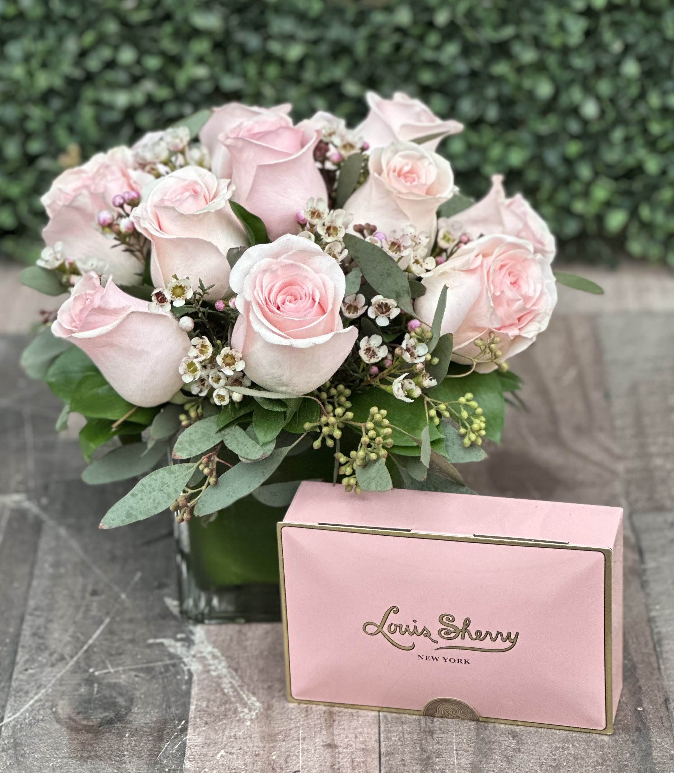 CLUSTER OF LOVE (LIGHT PINK) WITH LOUIS SHERRY PREMIUM CHOCOLATES - This clustered arrangement comes designed with roses, seeded eucalyptus, wax flower and assorted greenery in a cube wrapped with a leaf. It is paired with a Valentine's Teddy bear and a heart shaped artisan chocolate. Bear and chocolate come in a decorative window bag.