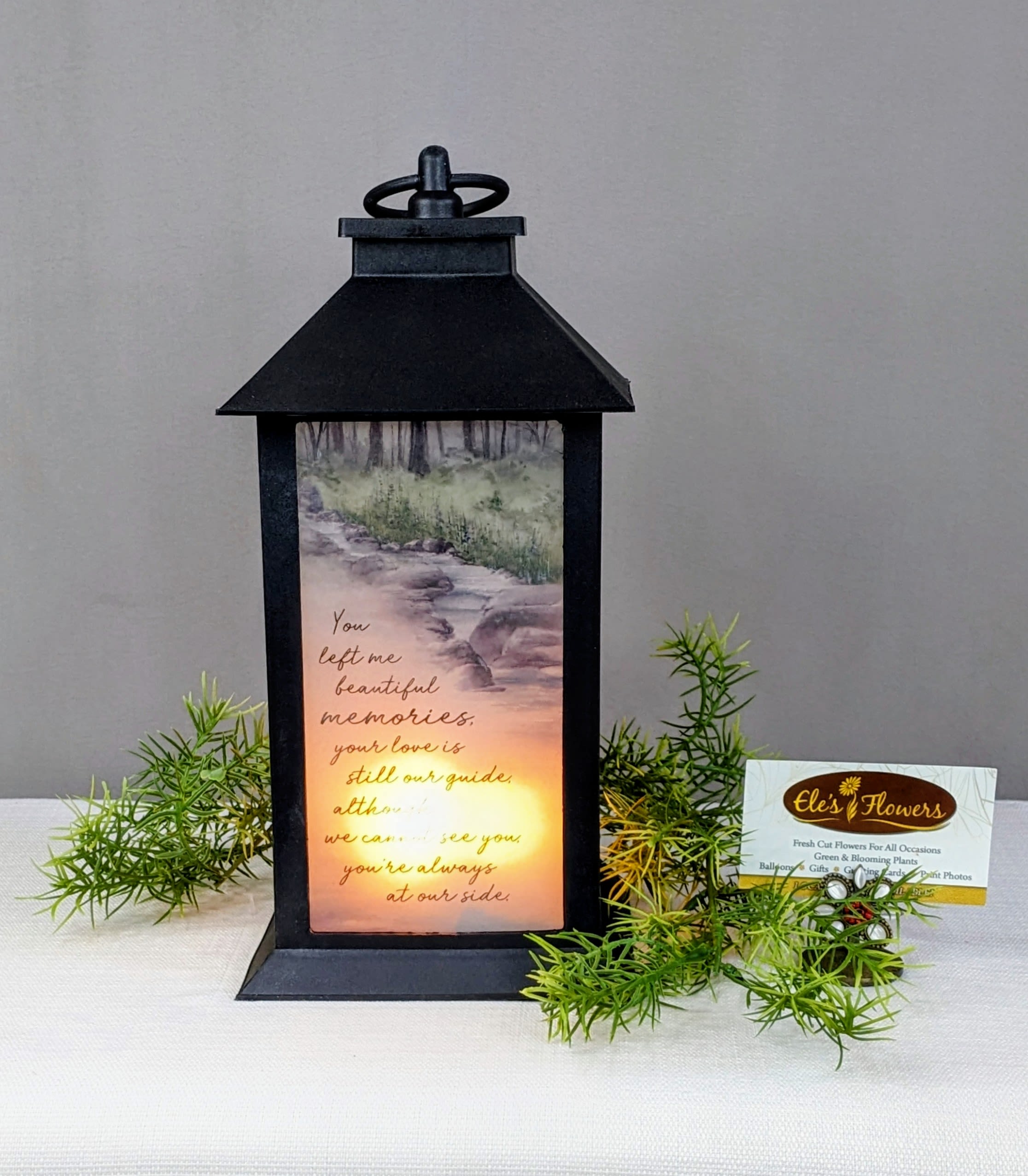 Black memorial lantern with glowing light and verse, accented by small green sprigs.