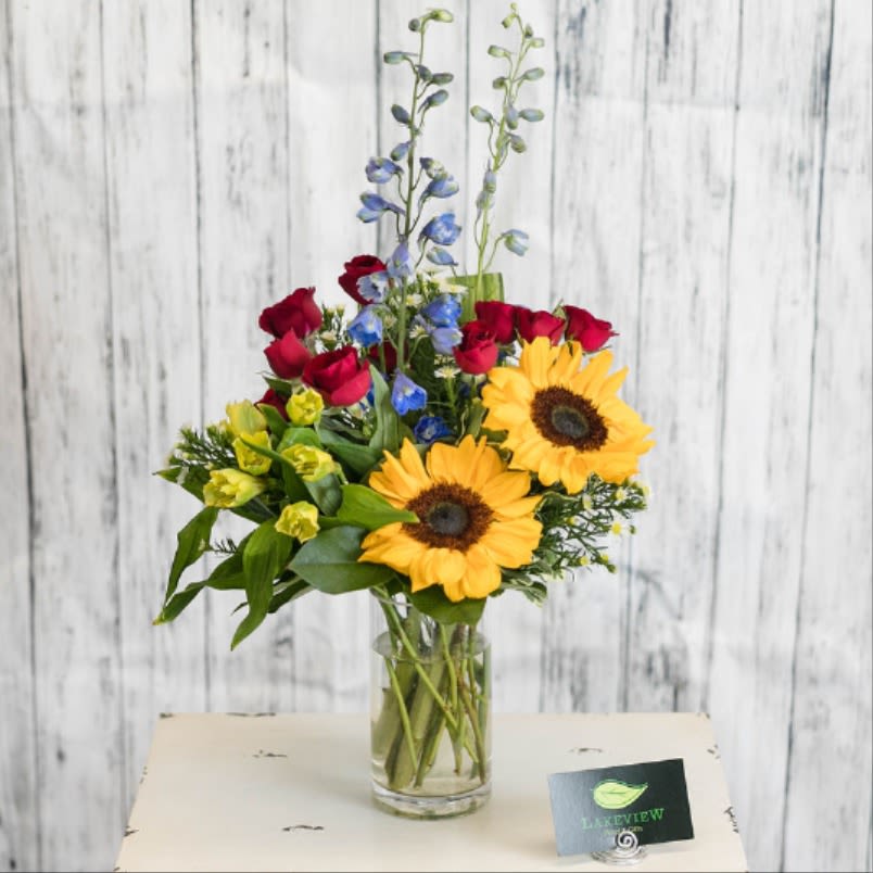 Grandstand  - Classic and strong colors like red spray roses, yellow sunflowers and blue delphinium, in a simple glass vase are sure to turn heads anywhere.   Approximate Dimensions: 22"H x 12"W