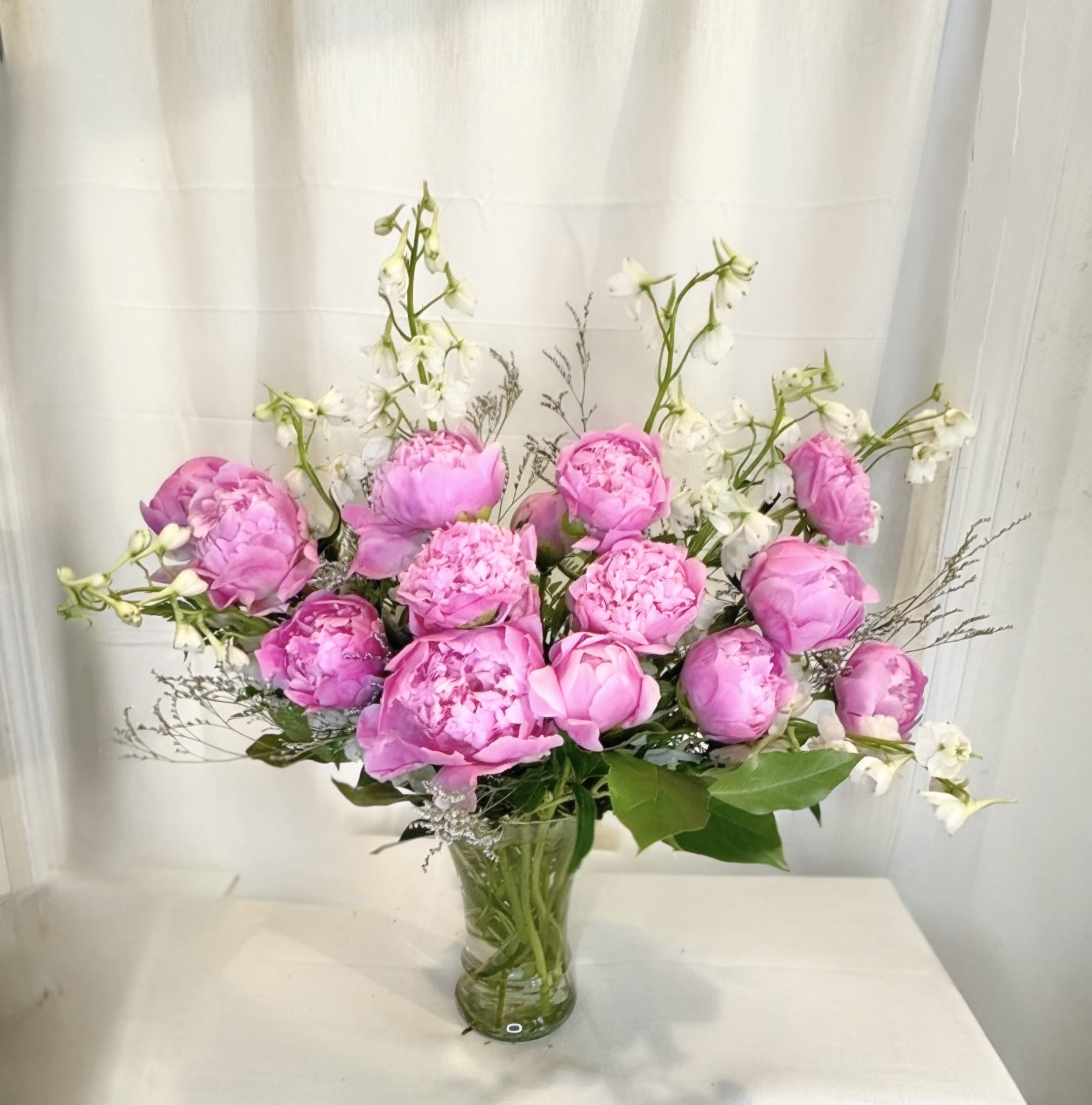 * Pink Peony Club * - A lush cluster of soft pink peonies takes center stage, surrounded by airy white accents that lift the composition and add a light, springtime feel. Designed to make the person you're thinking of feel cherished, the mix of romantic pinks and crisp white creates a cheerful yet elegant impression perfect for spring gifting or any moment that calls for celebration.  Peonies and complementary white filler blooms are gathered densely and set in a clear glass vase, the visible stems giving the design a fresh, garden-picked look. The rounded, full peony heads provide plush texture while the taller white accents add movement and contrast; colors are pink, white, and green for a classic, feminine palette that reads beautifully on a table or mantle.   The clear vase is ready to display on arrival; keep stems trimmed and change the water every other day to prolong the life of the peonies.