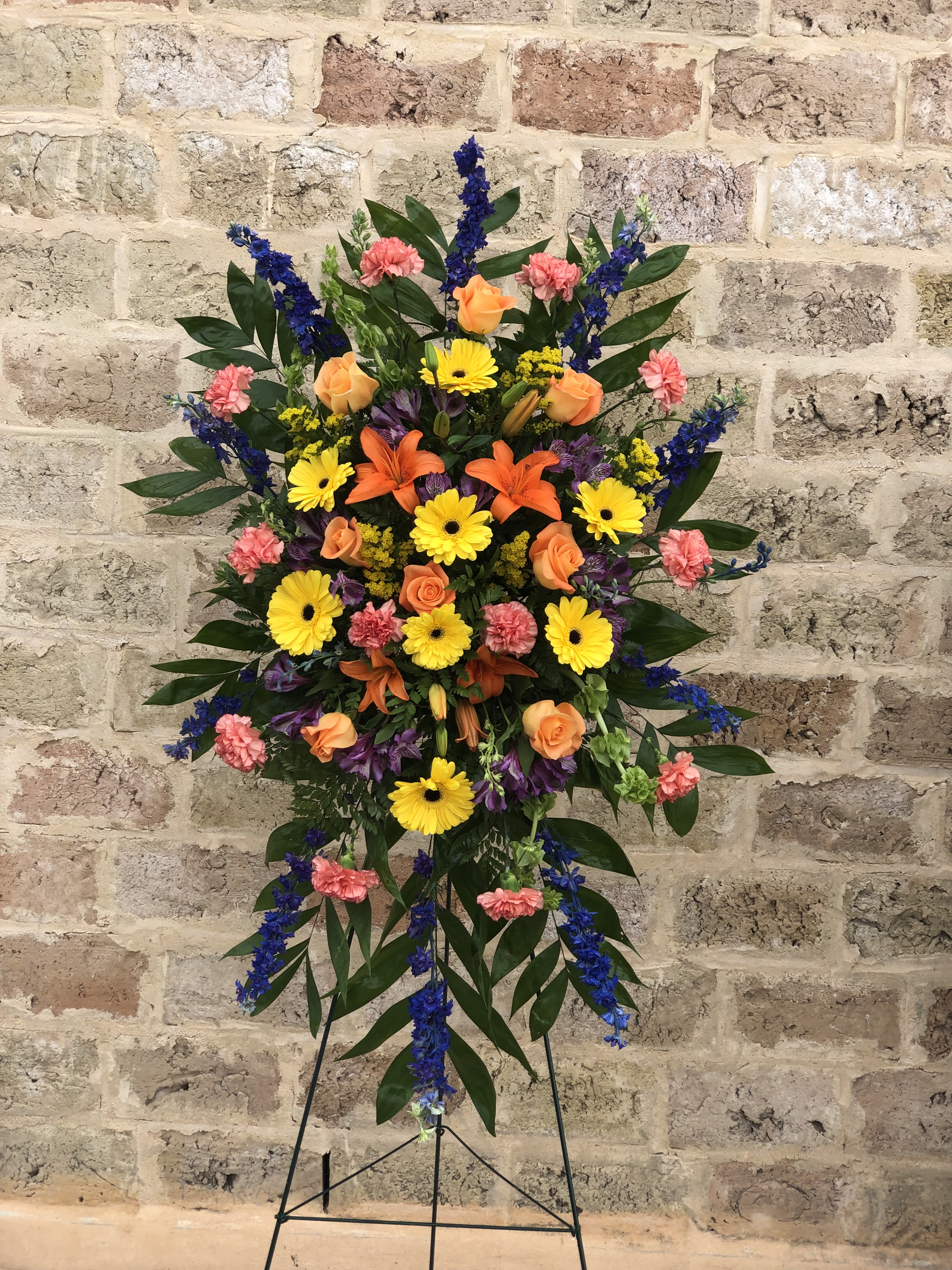 Standing floral spray with orange roses, yellow daisies, and blue accents