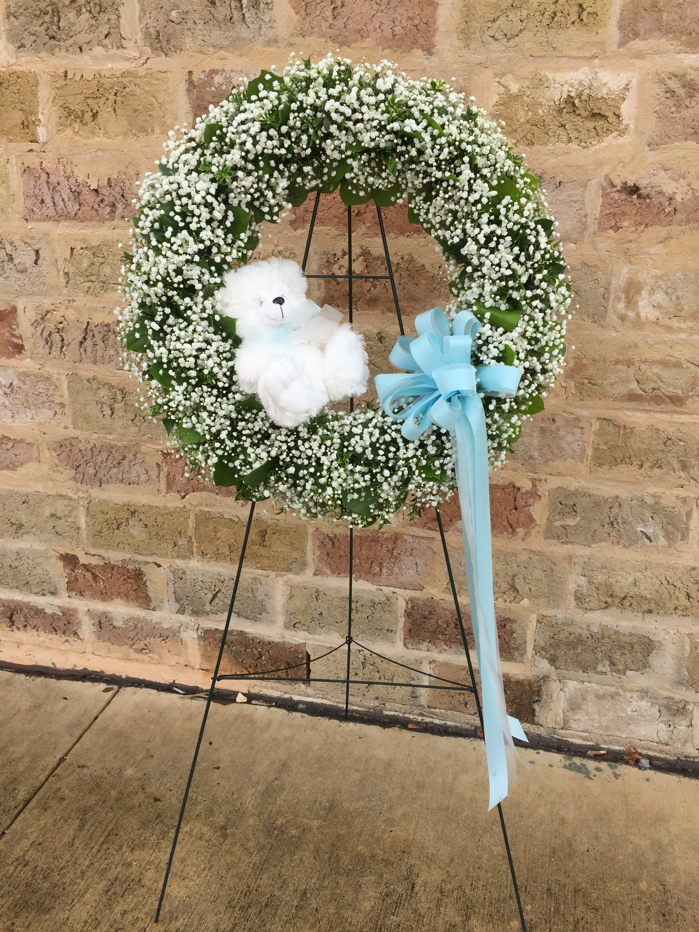 White baby’s breath wreath with white teddy bear and blue bow on a stand against a brick wall
