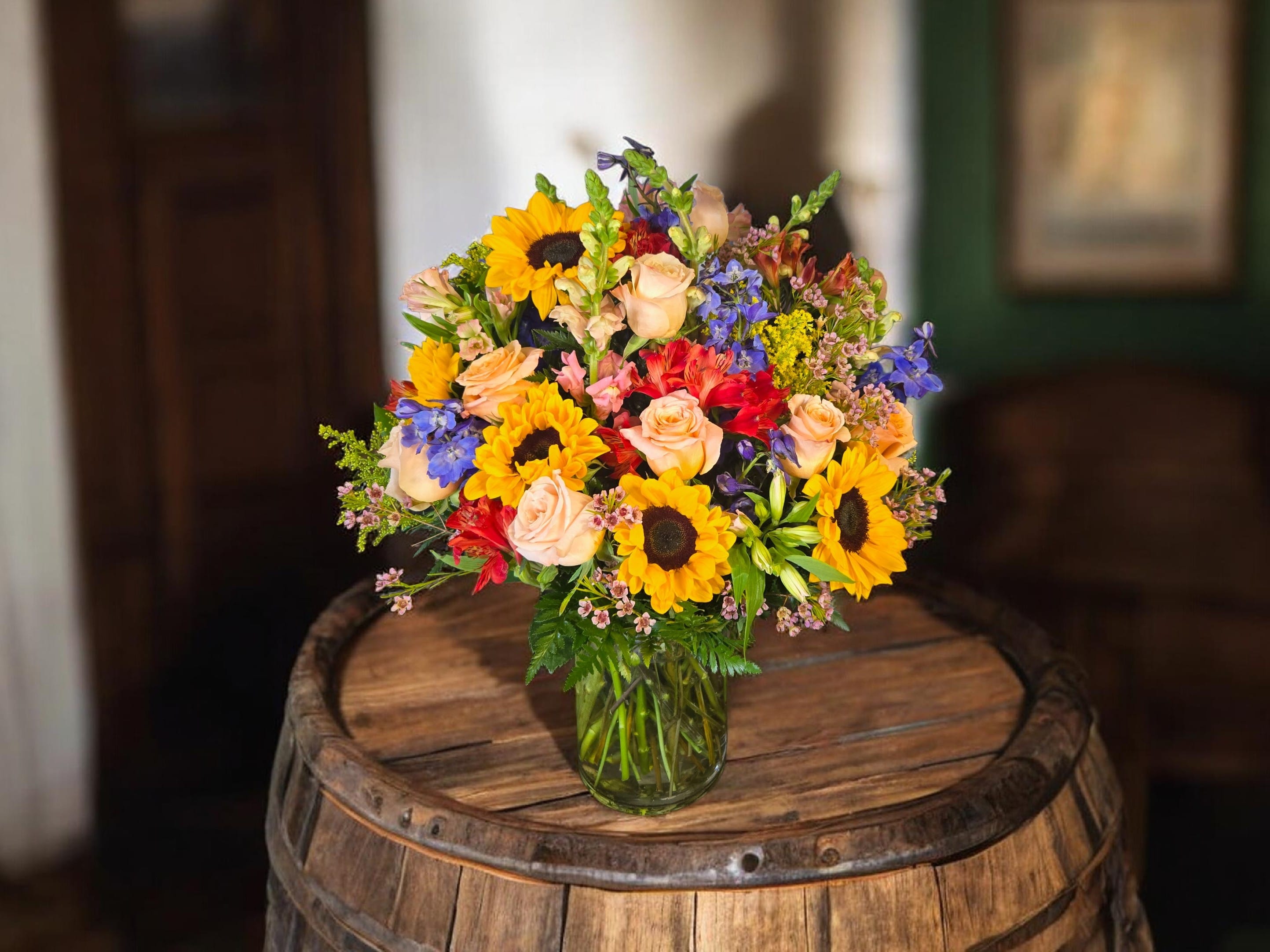 Sunlit Garden Bliss - Bright yellow sunflowers take center stage in this cheerful flower arrangement, a sunlit garden-style bouquet perfect for a birthday bouquet, congratulations, or just to brighten someone's day. The mix of sunflowers, blush roses, red blooms and small blue accent flowers creates an upbeat, welcoming look that reads as celebratory and genuinely joyful-the kind of gift that makes the person you're thinking of feel seen and smiled at the moment they walk in the room.  Arranged in a clear glass vase, the composition is full and rounded with a dense, layered feel that suggests casual elegance. Blush roses soften the bold sunflower heads while red blooms add lively contrast and the cool blue accents provide a subtle pop that balances the palette. The texture is varied-broad sunflower faces against the ruffled roses and dainty alstroemeria-so the bouquet reads as abundant without feeling fussy.  