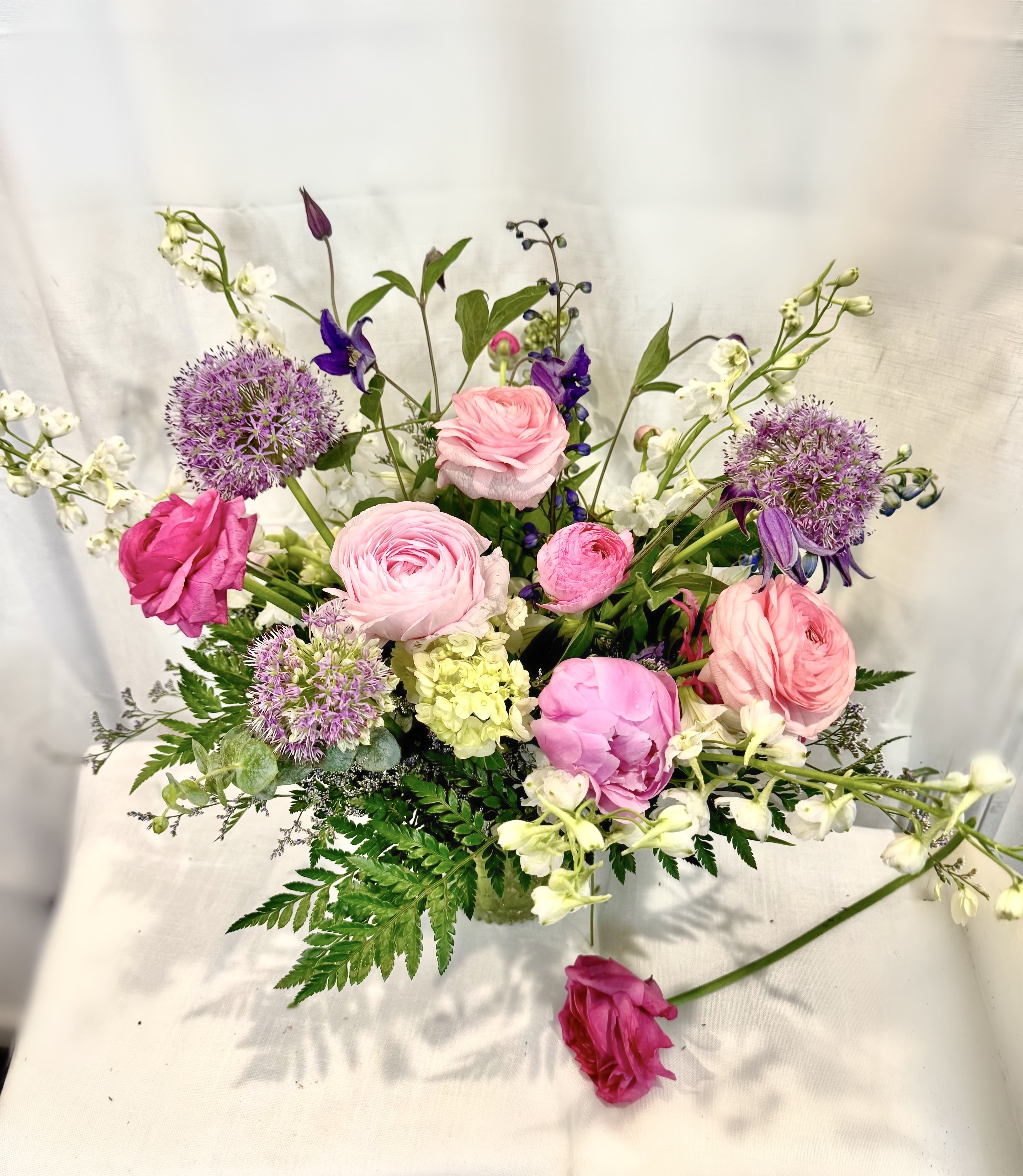 * Mom's Garden * - Layered pink ranunculus and roses mingle with tall blue‑purple delphinium and pops of spherical purple allium in a loose, garden‑style flower arrangement perfect for a birthday bouquet, spring celebration, or a heartfelt Mother's Day gift. The soft pinks and whites read romantic and feminine while vivid violet accents lift the composition-she'll feel noticed and genuinely celebrated the moment this arrives.  This cheerful arrangement pairs delicate white alstroemeria and abundant greenery with varied textures-round blooms, spiky delphinium, and branching filler-for an airy, asymmetrical silhouette that looks freshly gathered from a cottage garden. Arranged in a clear glass vase, the presentation stays light and natural so it brightens a kitchen table, bedside, or office desk without overwhelming the space.  Ideal for moms, friends, or anyone who loves soft pastels with a lively twist, this bouquet sends a message of warmth and appreciation for everyday moments or special springtime occasions. Care is simple: keep the water fresh and display in bright, indirect light to enjoy the flowers at their best.