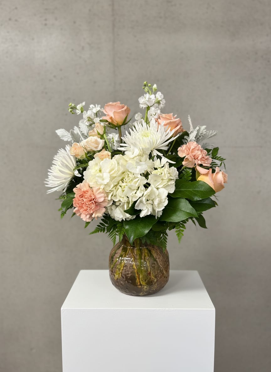 Honey Glow - Honey Glow feels like a warm, late-afternoon light captured in flowers. Soft peach tones from the roses and carnations give the arrangement its gentle radiance, while delicate pink spray roses add a subtle blush that keeps it from feeling too uniform. Clouds of white hydrangea and airy white stock bring fullness and a sense of calm, balanced by the unique texture of white spider mums that stretch outward like quiet bursts of light.  Dusty Miller weaves through it all with its silvery, velvety leaves, adding a muted contrast that softens the sweetness and gives the piece a slightly vintage feel. Arranged in a peach-toned vase, everything comes together in a way that feels both glowing and serene—like honeyed sunlight resting on a windowsill.