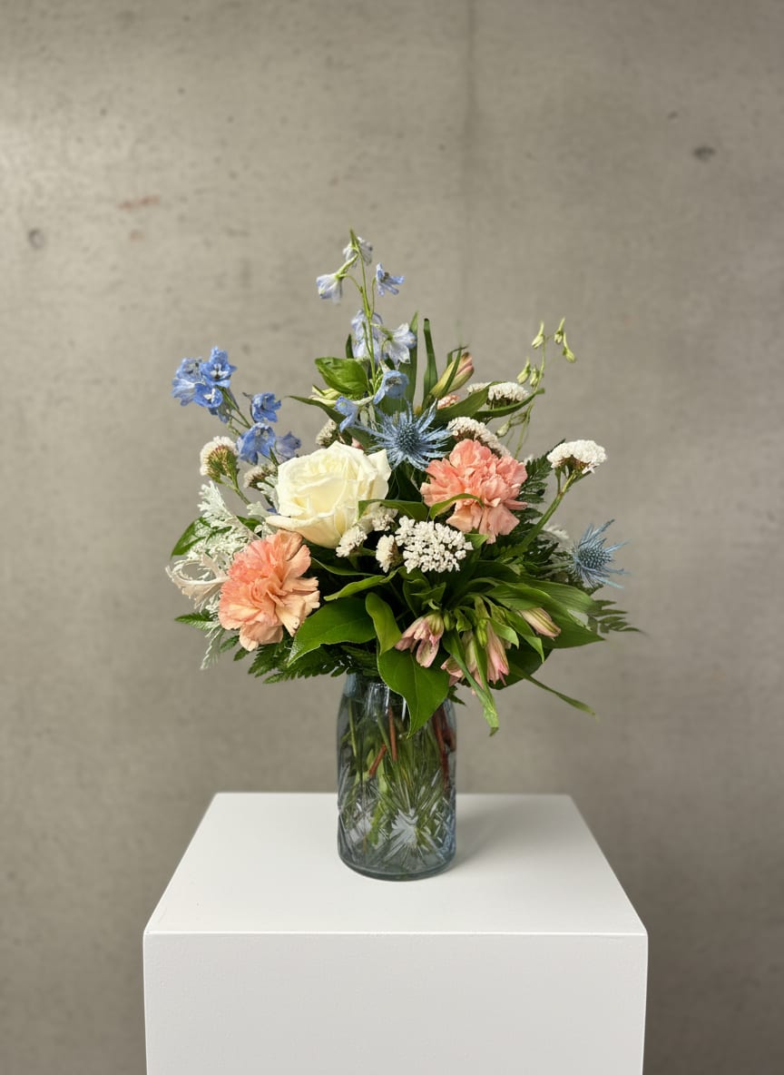 Garden Grace - Garden Grace is a soft, airy arrangement that feels like a peaceful morning in bloom. Set in a sky blue vase, it features classic white roses alongside pink carnations and delicate pink alstroemeria for a gentle, romantic palette. Light blue delphinium rises above the arrangement, adding height and a touch of sky-like serenity, while hints of thistle introduce texture and a subtle wild beauty. Dusty miller weaves in silvery softness, and white statice provides a light, lasting finish—bringing everything together in a design that feels both graceful and naturally elegant.