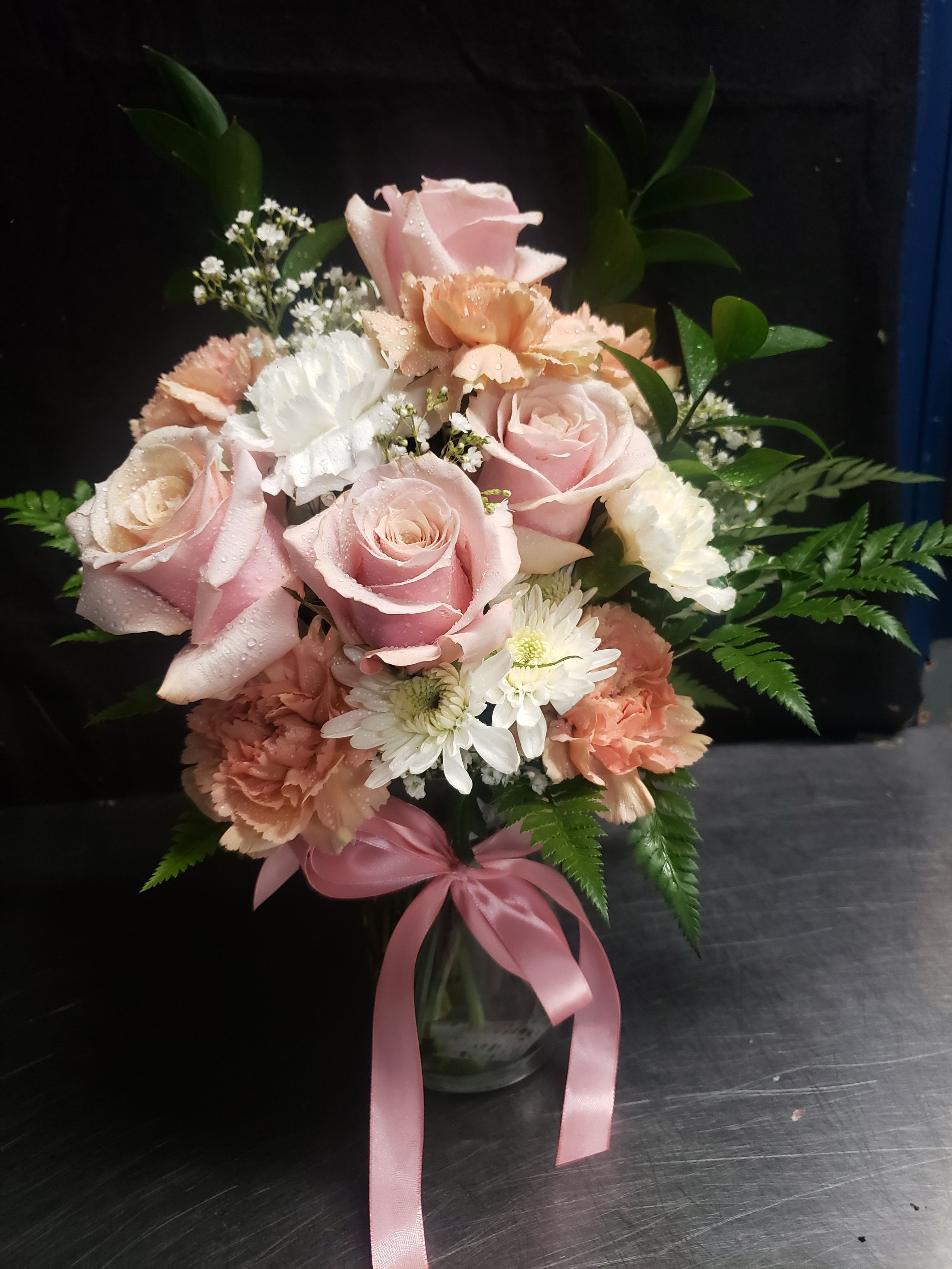 Love Bouquet with pink - Soft pink roses and peach-and-white carnations mingle with tiny white daisy-like blooms in a gentle, romantic bouquet-perfect as a birthday bouquet, anniversary arrangement, or a thoughtful Mother's Day gift. The pastel palette and rounded, full shape read as fresh and feminine; the person you're sending it to will feel adored and seen the moment this blooms-filled vase arrives.  Arranged in a clear glass vase and finished with a pink satin ribbon, this floral arrangement includes airy filler flowers and fern-like foliage for texture and movement that keeps the design lush but compact. Colors: pink, white, peach, and green. Flowers: roses, carnations, daisies, plus additional filler for a hand-arranged, classic presentation.  Ideal for everyday gifting, saying "thank you," or marking a special milestone, this bouquet arrives ready to display-no extra work required. For best results, place in bright, indirect light, refresh the water every couple of days, and trim stems slightly when you change the water to prolong vase life.