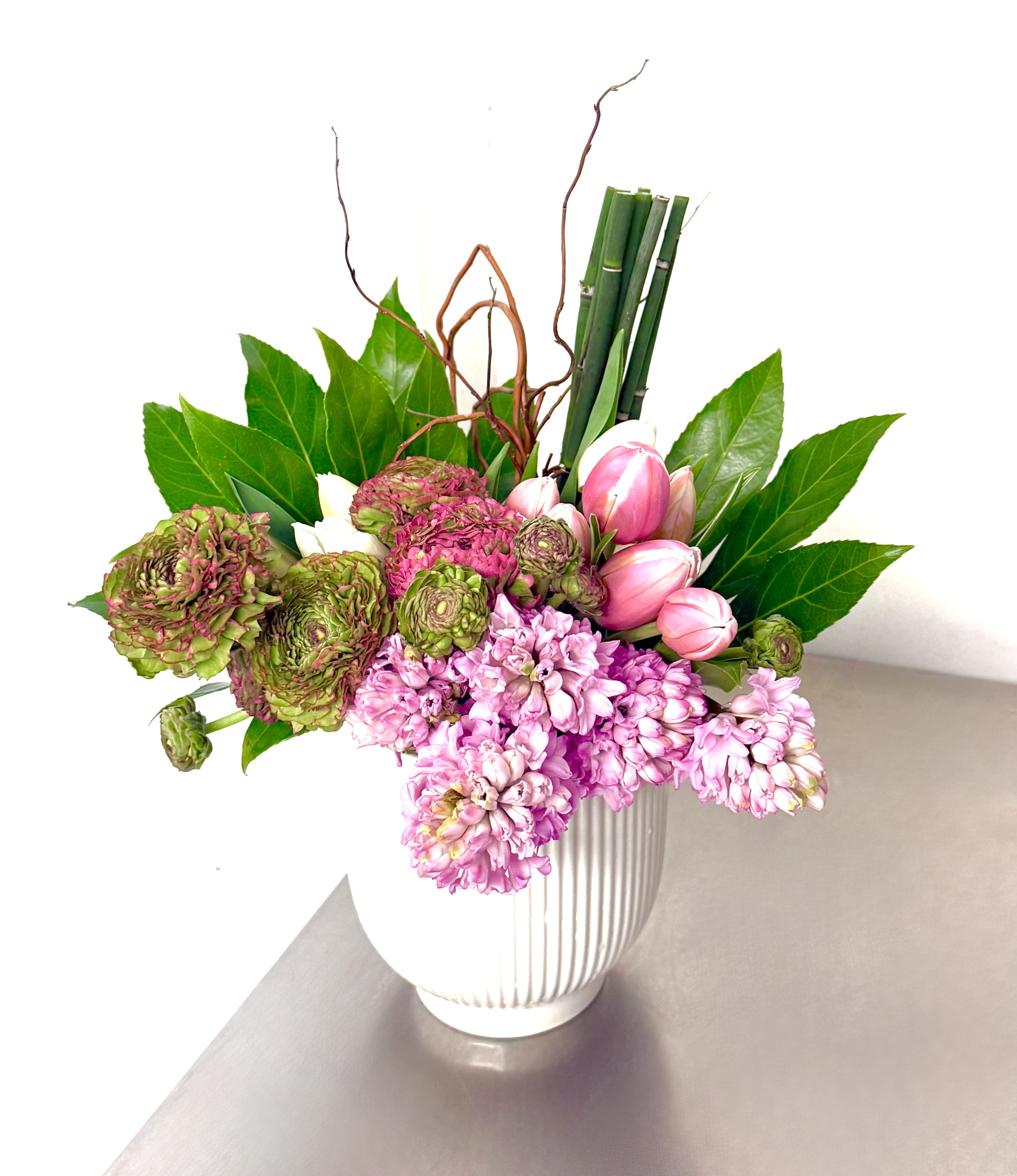Garden Serenade - Bring the vibrant energy of spring to your home with our Garden Serenade arrangement. This exquisite bouquet features a harmonious mix of premium blooms, showcasing the best of the season's textures and scents. The Blooms: Delicate, ruffled pink ranunculus and whimsical tulips provide soft, romantic layers. Fragrant pink hyacinth clusters ground the design with their intoxicating spring perfume and rich, detailed florets. The Details: Accented by striking bamboo stalks and twisting natural branches for architectural height, and framed by glossy, deep green foliage for a lush, full-bodied look. 