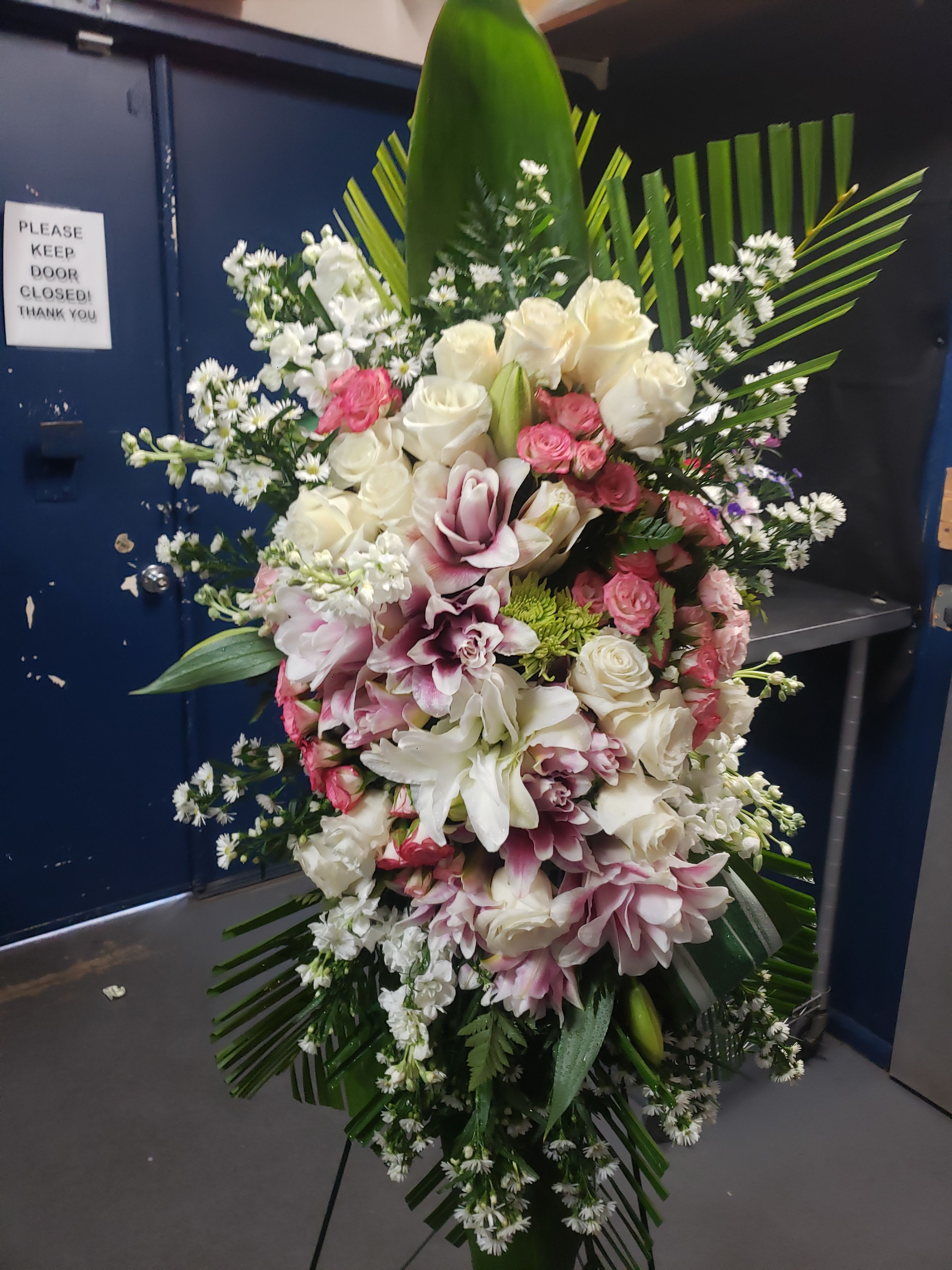 Funeral, Bouquets and Arrangement's - A tall, graceful standing spray of soft pink roses, white lilies, and delicate daisy-like blooms creates a gentle, comforting tribute ideal for a sympathy arrangement or memorial service. The vertical, layered design and white ribbon streamers give this funeral spray a formal, reverent presence that will make the person you're honoring feel remembered and respected.  This spray is built with pale pink roses at the heart, surrounded by white lilies, pink carnations, hydrangea clusters, and small white filler blooms for an airy texture. Broad dark green foliage and palm fronds form a dignified backdrop while the layered composition on an easel ensures the arrangement reads clearly from a distance. The overall palette of soft pinks and whites keeps the look tender and soothing.  Appropriate for a funeral, remembrance service, or as a consoling delivery to family, this piece communicates sympathy and gentle support. Sizes and installation on a metal easel are suited for display at a church or memorial site; please indicate any personalization for ribbons or messaging when ordering to ensure your tribute is presented exactly as you intend.