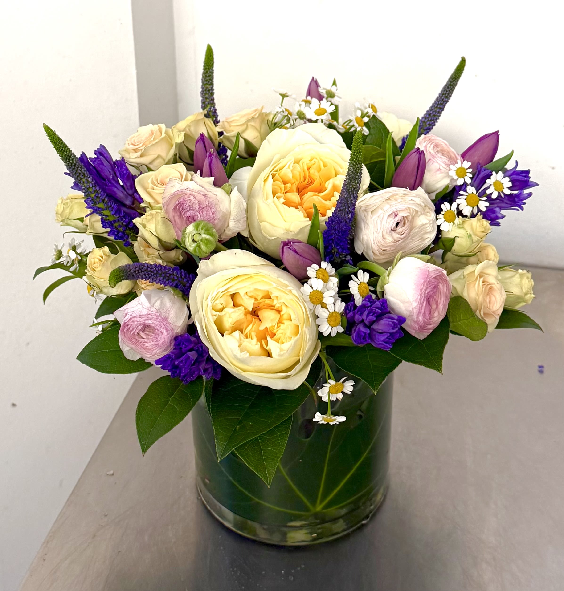 Luminous Garden Reverie - Experience the charm of a sun-drenched meadow. This enchanting design features buttery-yellow garden roses and ethereal ranunculus nestled among playful purple tulips and cheerful chamomile. Spikes of deep violet veronica add a touch of untamed elegance, all beautifully presented in a leaf-wrapped cylinder vase for a sophisticated, "straight-from-the-garden"