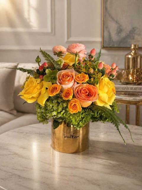 Golden Sunset - A radiant blend of golden yellows, warm peaches, and soft coral tones, this arrangement captures the glow of a perfect sunset. Designed with lush roses, delicate ranunculus, and fresh greenery, it brings a refined yet joyful presence to any space. Elegant, warm, and effortlessly timeless.   APPROXIMATE DIMENSIONS: 12" H X 10" W X 10"L