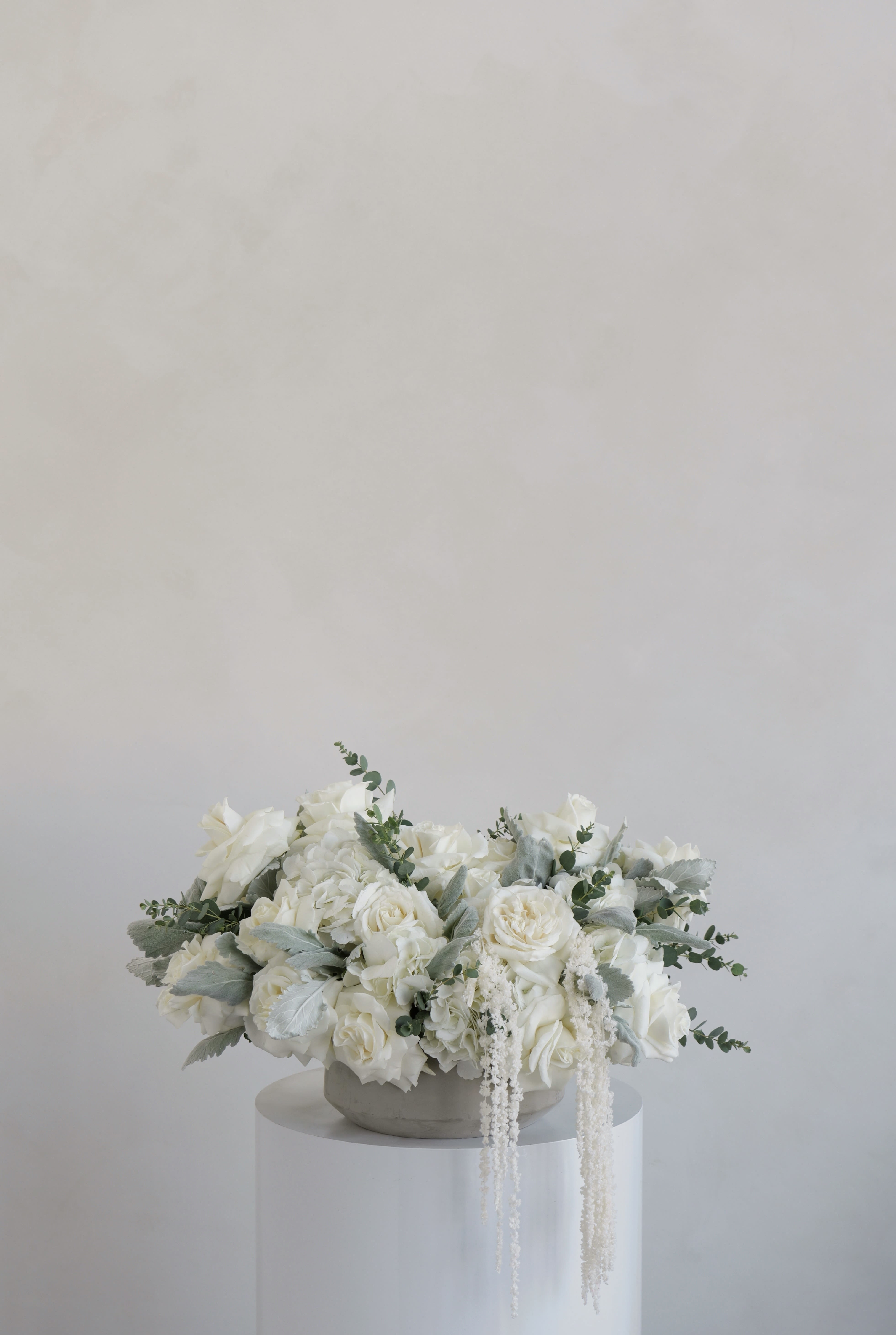 Evelina - A timeless, elegant arrangement designed in soft neutral tones, featuring lush white blooms paired with delicate greenery. The composition is full and balanced, with gentle cascading accents that add movement and texture. Designed in a low, modern vessel, this piece creates a refined and sophisticated look, perfect for any occasion. 