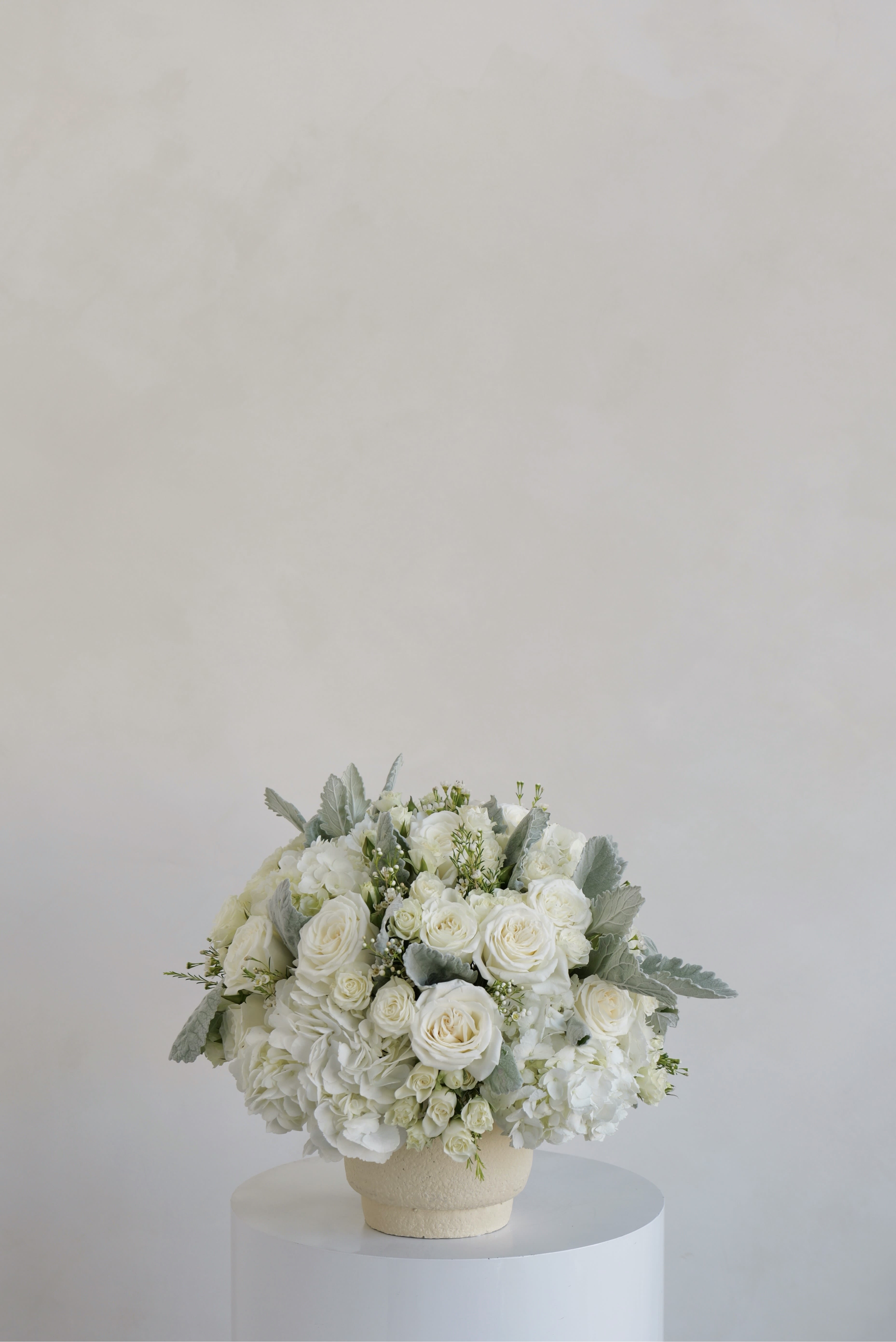 Elena - A classic, full-bodied arrangement featuring lush white blooms thoughtfully gathered into a soft, rounded silhouette. Layers of roses and hydrangea create rich texture and depth, while subtle greenery adds a natural, airy touch. Designed in a low, textured vessel, this piece offers a clean and timeless look, perfect for elevating everyday spaces or adding understated elegance to special occasions. 