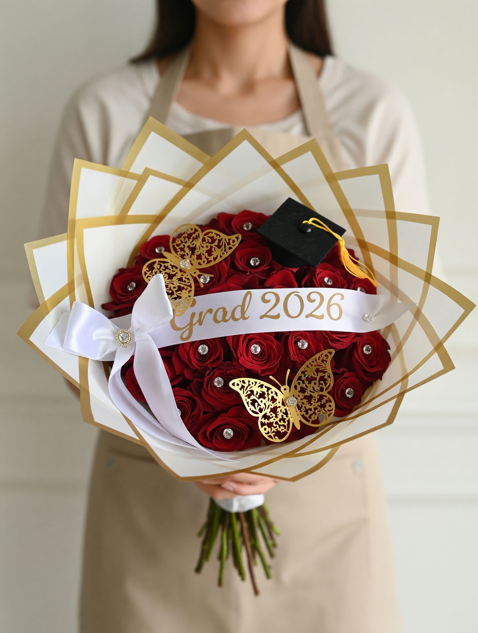 Graduation Bouquet 2026 / Ramo de Graduación 2026 - Graduation Bouquet 2026 / Ramo de Graduación 2026  Celebrate success in style with our Graduation Bouquet 2026, featuring luxurious red roses adorned with crystal accents, golden butterflies, and a mini graduation cap. Wrapped in elegant white and gold paper with a custom “Grad 2026” ribbon, this bouquet is the perfect way to honor hard work and new beginnings.  Celebra el éxito con estilo con nuestro Ramo de Graduación 2026, elaborado con elegantes rosas rojas decoradas con cristales, mariposas doradas y un pequeño birrete de graduación. Envuelto en papel blanco y dorado con cinta personalizada “Grad 2026”, es el detalle ideal para reconocer logros y nuevos comienzos.  Perfect for / Perfecto para: Graduation celebrations and milestone achievements. Includes / Incluye: Fresh red roses, gold and white wrapping, butterflies, crystal accents, and ribbon. Note / Nota: Custom text available upon request.