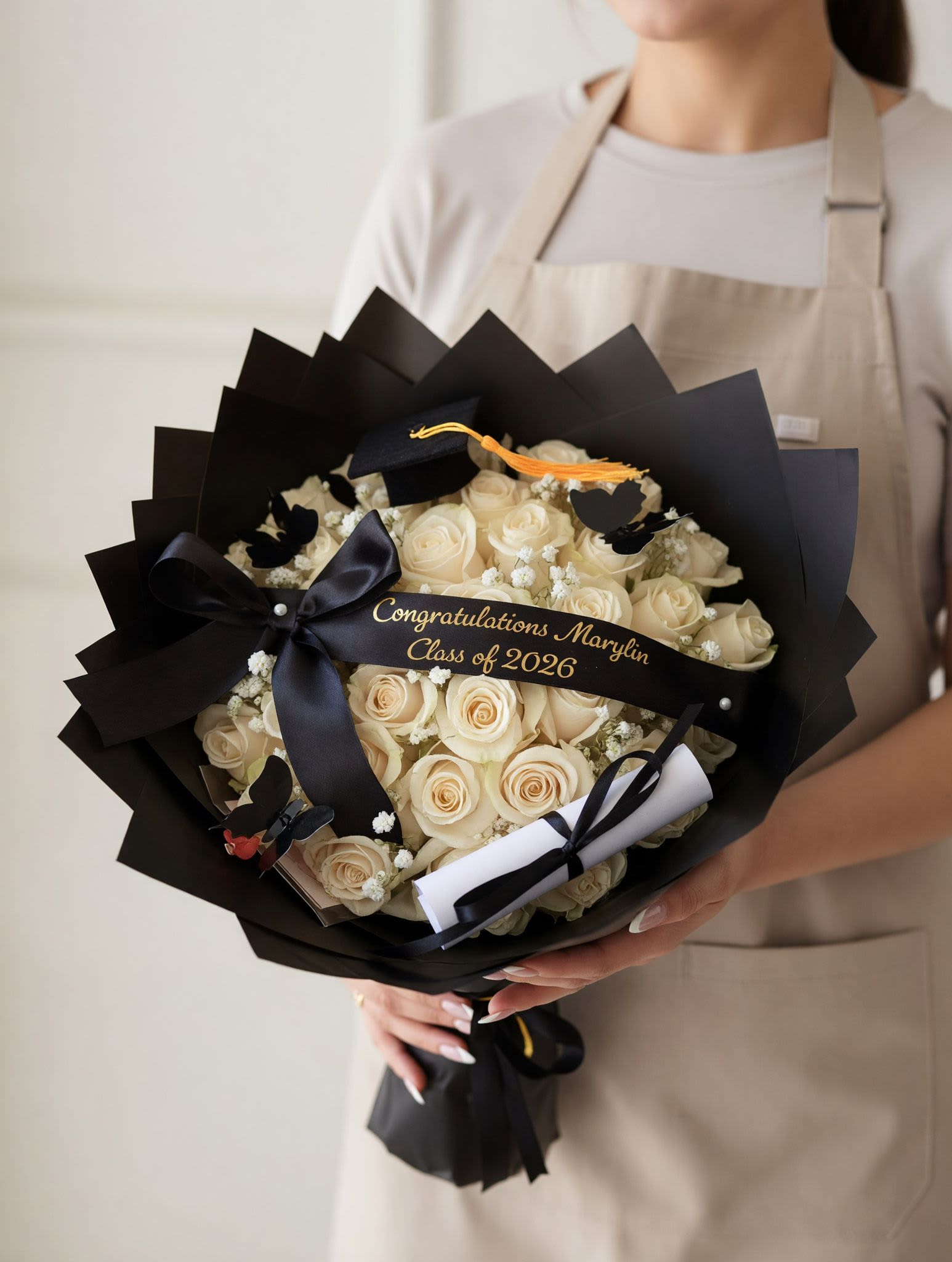 Class of 2026 Graduation Bouquet - Celebrate their milestone in the most unforgettable way with our Graduation Luxe Rose Bouquet. This elegant arrangement features premium fresh roses beautifully wrapped in sleek black paper and finished with a satin bow for a sophisticated touch. Accented with graduation details like a mini cap and personalized message ribbon, it’s the perfect gift to honor their hard work and success.  Designed to impress and made with love, this bouquet is ideal for graduations, achievements, and special moments worth celebrating. Each arrangement is handcrafted and may vary slightly to ensure the freshest and most beautiful presentation.