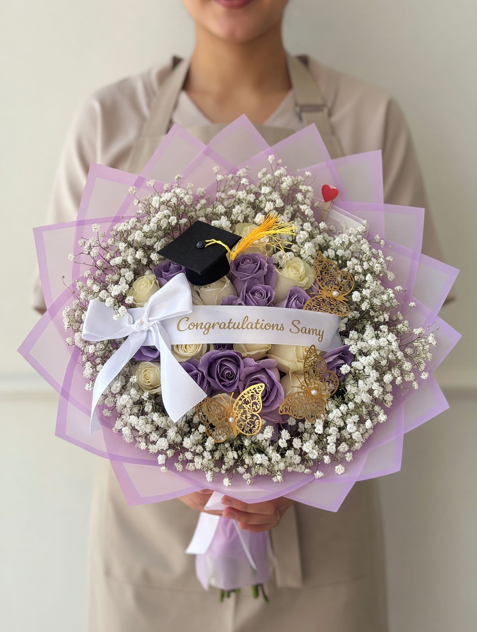 White &amp; Lilac Graduation Bouquet - Celebrate their milestone in the most delicate and elegant way with our Lilac Elegance Graduation Bouquet. This handcrafted design features a soft blend of lavender and ivory roses, beautifully surrounded by airy baby’s breath for a full, cloud-like effect.  Wrapped in layered lilac-toned paper and finished with a satin bow, this bouquet is accented with thoughtful graduation details including a mini cap, gold butterfly accents, and a personalized message ribbon—creating a truly meaningful and memorable gift.  Perfect for graduations and special achievements, this bouquet is designed to stand out while maintaining a soft, feminine aesthetic. Each arrangement is made fresh to order using premium blooms.  Customization available for name, ribbon message, and color tones.
