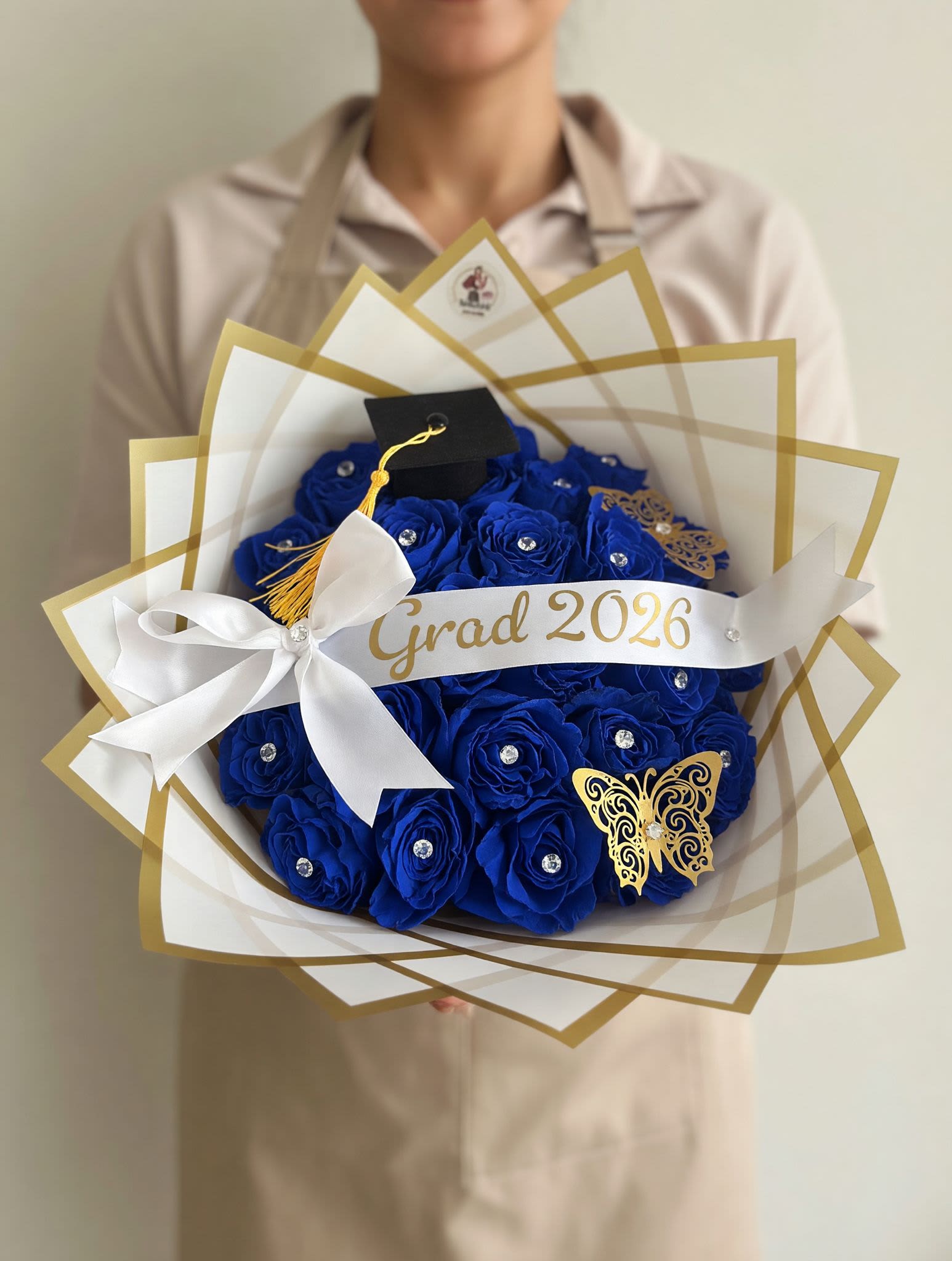 Grad 2026 Graduation Bouquet Blue Roses - Graduation Bouquet 2026 / Ramo de Graduación 2026  Celebrate success in style with our Graduation Bouquet 2026, featuring luxurious blue roses adorned with crystal accents, golden butterflies, and a mini graduation cap. Wrapped in elegant white and gold paper with a custom “Grad 2026” ribbon, this bouquet is the perfect way to honor hard work and new beginnings.  Celebra el éxito con estilo con nuestro Ramo de Graduación 2026, elaborado con elegantes rosas azules decoradas con cristales, mariposas doradas y un pequeño birrete de graduación. Envuelto en papel blanco y dorado con cinta personalizada “Grad 2026”, es el detalle ideal para reconocer logros y nuevos comienzos.  Perfect for / Perfecto para: Graduation celebrations and milestone achievements. Includes / Incluye: Fresh red roses, gold and white wrapping, butterflies, crystal accents, and ribbon.