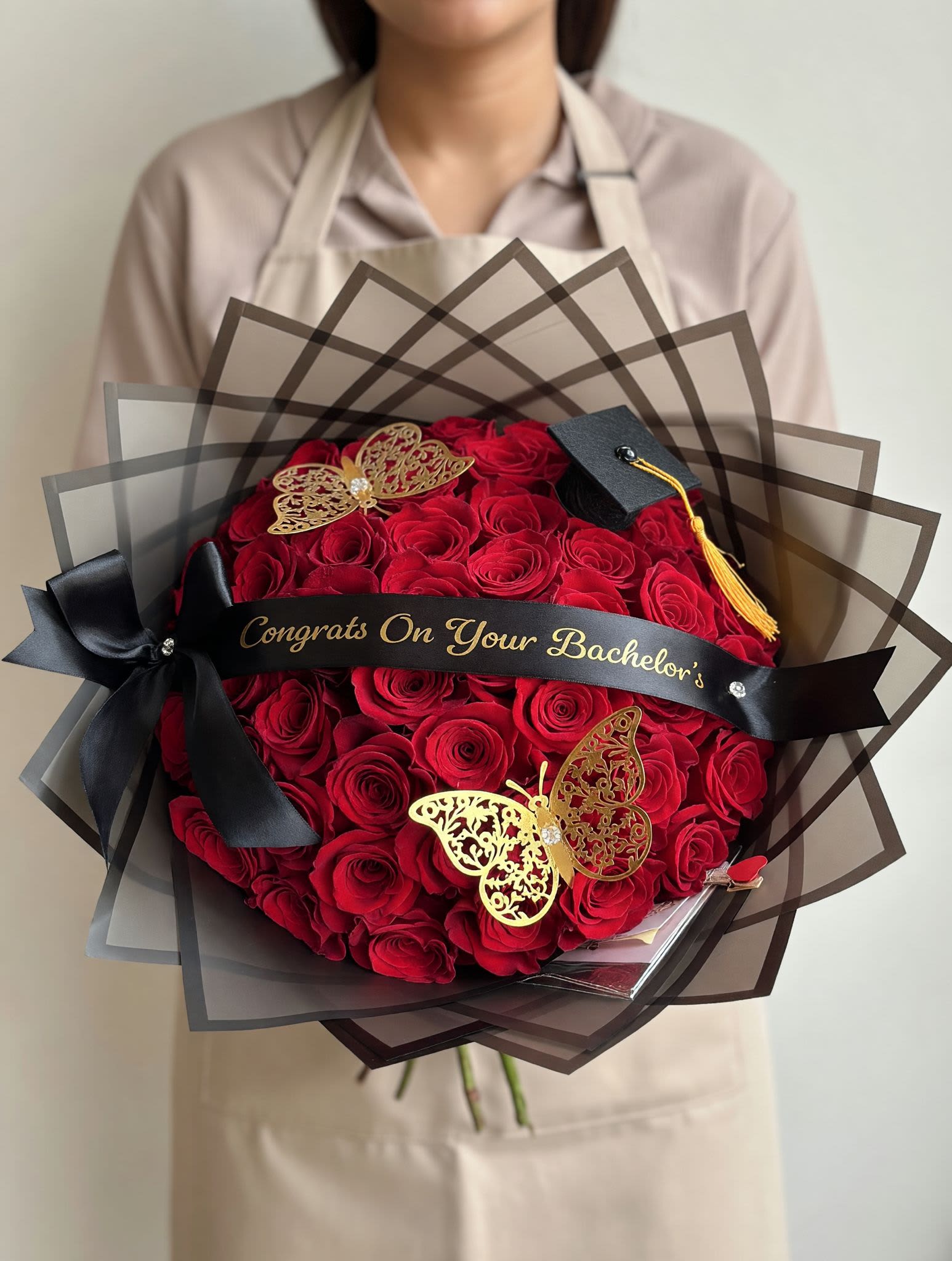 50 Roses Congrats On Your Bachelor's Graduation Bouquet Custom Ribbon - Make their achievement unforgettable with our Midnight Red Graduation Bouquet — a bold and luxurious design crafted to celebrate life’s biggest milestones.  This stunning bouquet features rich, velvety red roses arranged in a full, rounded style and wrapped in layered black translucent paper for a modern, high-end look. Finished with a satin ribbon, gold butterfly accents, and a graduation cap detail, this piece is designed to make a statement.  A personalized message ribbon adds the perfect finishing touch, making this bouquet both meaningful and memorable. Ideal for graduations, promotions, and special celebrations.  Each bouquet is handcrafted fresh to order using premium roses for maximum beauty and impact.  Customization available for ribbon message and accents.