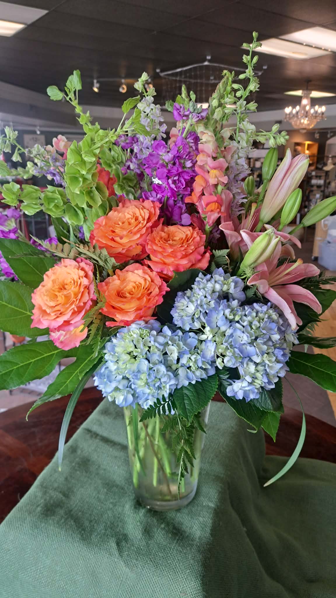  Devoted​ - Bright, garden-fresh blooms arranged in a clear glass vase-this mixed flower arrangement brings together tulips, hydrangeas, lilies, snapdragons, roses and seasonal fillers for a cheerful, springlike gift. As a birthday bouquet, get-well gesture, or thank-you arrangement, it reads as both uplifting and composed: tall yellow tulips and purple spikes give vertical energy while rounded blue hydrangea heads and lush green foliage add calm fullness so the person you're thinking of feels seen and comforted.  Expect a lively mix of color and texture-pale blue hydrangeas, sunny yellow tulips and small yellow filler flowers for brightness; pink-and-white lilies and coral-orange roses for warmth; and upright purple and lavender accents for contrast. The arrangement arrives tucked into a clear glass vase and finished with a glossy purple satin ribbon for a polished, celebratory touch. Sizes are arranged to present layered height and generous volume, making this a standout centerpiece for birthdays, anniversaries, congratulations, or simply to brighten someone's day; light water changes and fresh-cut stems will keep the bouquet looking its best.