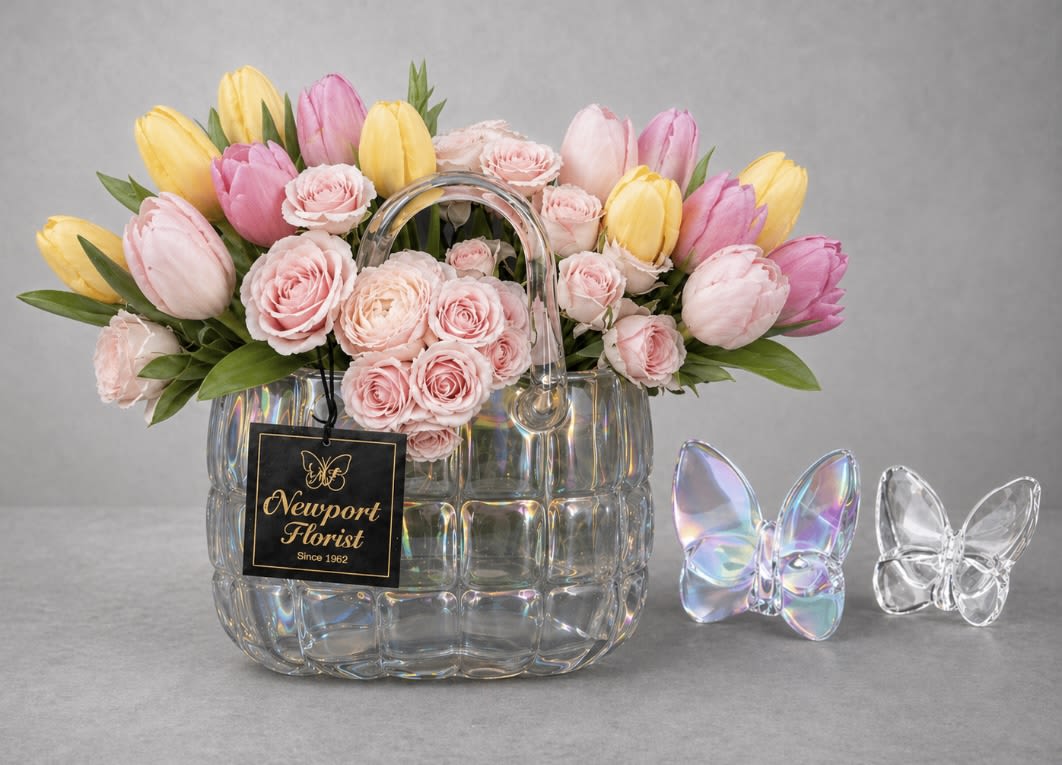 Pastel Tulip Garden in  Cyrstal handbag NF-Y42 - Pink and yellow tulips sweep in a fresh, springlike arc around a lush cluster of blush roses , making this flower arrangement an ideal birthday bouquet, spring gift, or light romantic gesture. The soft, cheerful palette of pinks, yellows, reads feminine and uplifting-perfect for the person you want to make smile.  Arranged in a clear, handbag-shaped glass vase, the blooms feel fashion-forward and playful; decorative butterfly ornaments add a whimsical touch and a florist tag finishes the look. The tulips provide graceful movement while the roses give the bouquet a full, textured center-an arrangement that looks effortless on a mantel, bedside table, or as a thoughtful surprise delivered to their door.