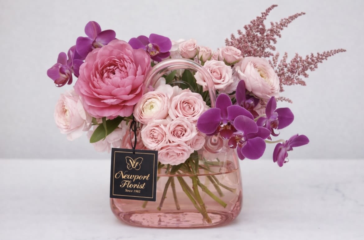 Pink Garden  Crystal Basket NF-Y43 - A lush, romantic bouquet built around a large pink ranunculus-style bloom and clusters of pale pink roses-perfect as a birthday bouquet or a celebratory gift. The inclusion of vivid purple Phalaenopsis orchids lifts the palette, with white accents and airy pink filler sprays giving the arrangement a soft, feminine silhouette that reads polished yet approachable for everyday gifting.  Arranged in a rounded pink glass vase with an attached floral tag, this design pairs blush and magenta tones for a refined contrast that catches the eye. The mix of ranunculus-like flowers, roses, orchids and delicate filler creates a variety of textures-velvety petals, waxy orchid blooms, and feathery sprays-so the recipient will feel truly celebrated. Ideal for anniversaries, congratulations, or simply saying "I'm thinking of you," available in a ready-to-display size that arrives gift-ready in its clear pink vessel.