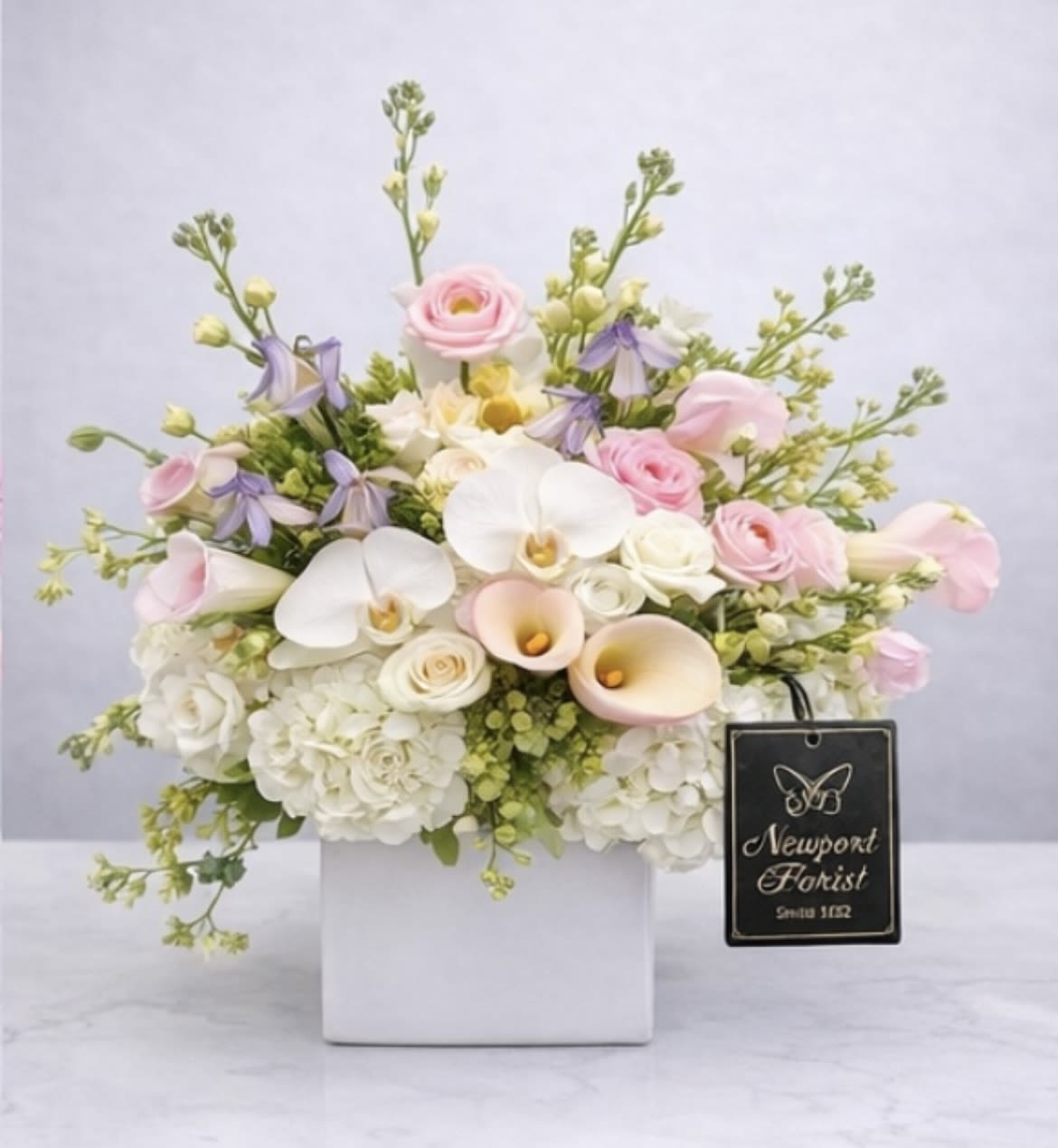 Aurora Blooms NF-Y46 - Soft pink roses and pale orchids create a gentle, romantic presence in this flower arrangement-perfect as an anniversary bouquet or an elegant everyday gift. The arrangement reads like a refined celebration: white calla lilies and creamy hydrangeas form a full, luxurious base while airy lavender accents and branching stems lift the composition, giving it a garden-fresh silhouette your recipient will notice.  Arranged compactly in a white ceramic square vase with a tasteful floral tag, the palette of white, pink, purple and touches of yellow feels both polished and soft. The mix of roses, calla lilies, hydrangeas and orchids offers a variety of textures-from the smooth curves of calla petals to the lush, cloud-like hydrangea heads-making the piece feel rich without being fussy. Sized to sit gracefully on a dining table or bedside, it's the kind of bouquet that makes someone feel truly seen and celebrated.  Ideal for marking anniversaries, weddings, or simply saying "I'm thinking of you," this arrangement arrives ready to display. Keep flowers in fresh water, out of direct sun and away from heat to extend their life; the ceramic vase and floral tag give it an upscale finish that's gift-ready the moment it's delivered.
