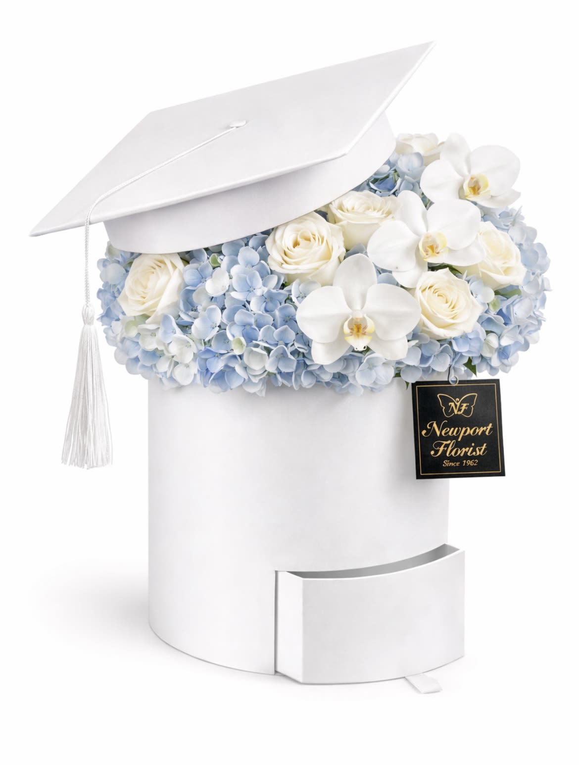 Cloud Nine Graduate NF-Y51 - Pale blue hydrangeas, white roses, and delicate white Phalaenopsis orchids spill from a white graduation cap-shaped box, creating a soft, celebratory centerpiece perfect for a graduation or congratulatory gift. This graduate flower arrangement is designed to make the recipient feel honored and seen-classic, calm pastels and refined textures give the impression of thoughtful achievement and milestone celebration.  The arrangement sits in a crisp white cap box with a coordinating cylindrical base and a real tassel, presented like a keepsake gift box. Tightly packed hydrangea blooms form a cloudlike cushion around clusters of cream-white roses and the elegant, sculptural orchids for a polished, photo-ready display. The compact, giftable size makes it easy to place on a desk, mantle, or dorm-room shelf as a lasting reminder of a proud moment.